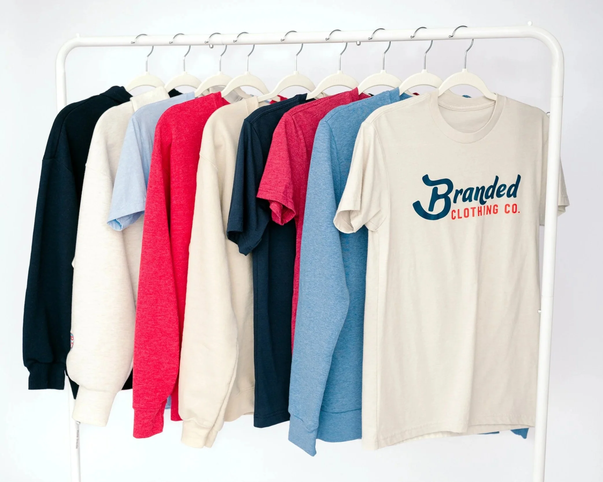 A clothing rack with nine T-shirts hanging on white hangers, featuring colors black, white, red, dark blue, light blue, and beige, with a beige T-shirt displaying a logo that reads 'Branded Clothing Co.'