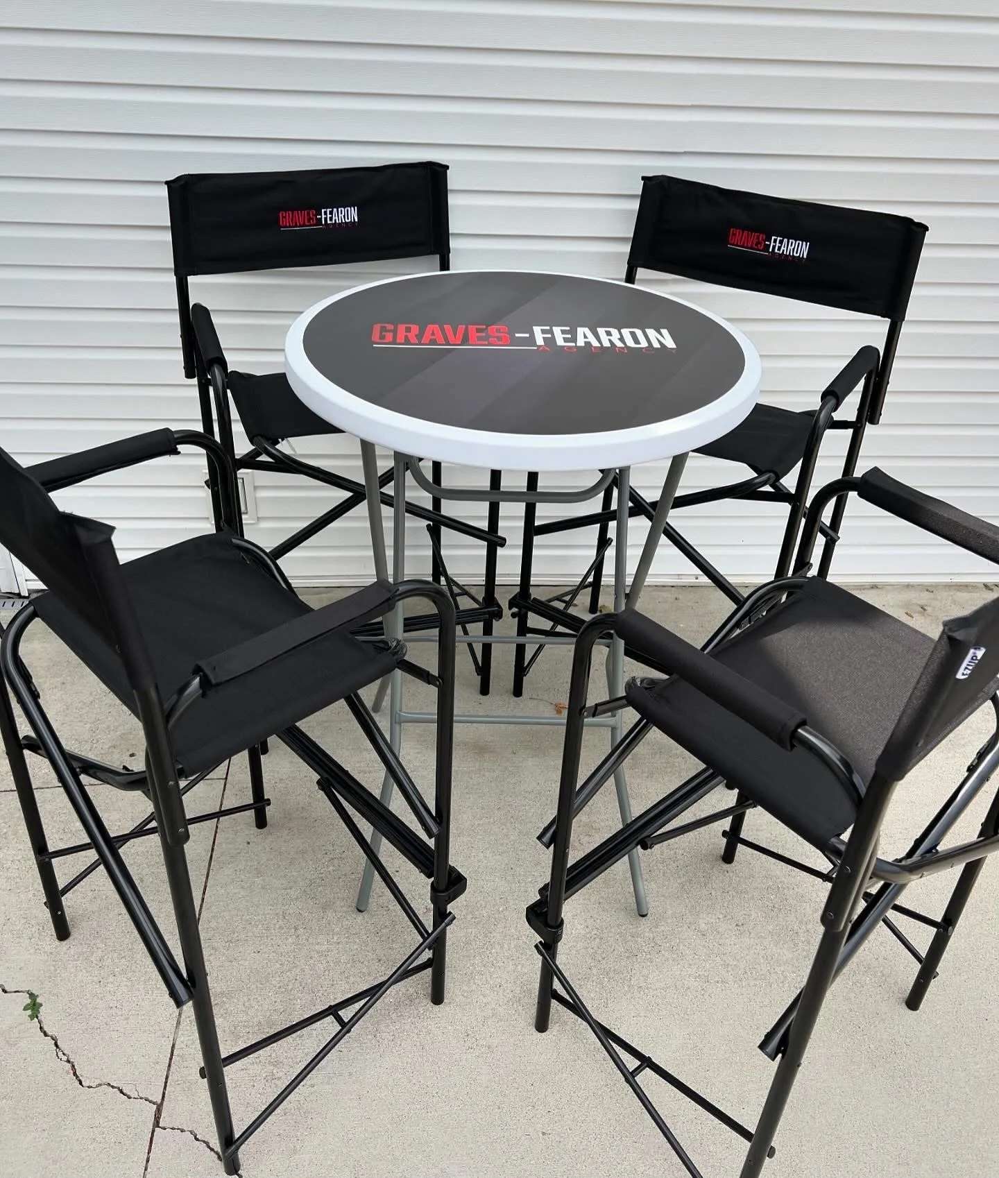 Outdoor patio table with four black folding chairs around it, with a white and black surface that has bold red and white text reading 'Graves-Fearon.' The chairs have black fabric backs with the same logo.