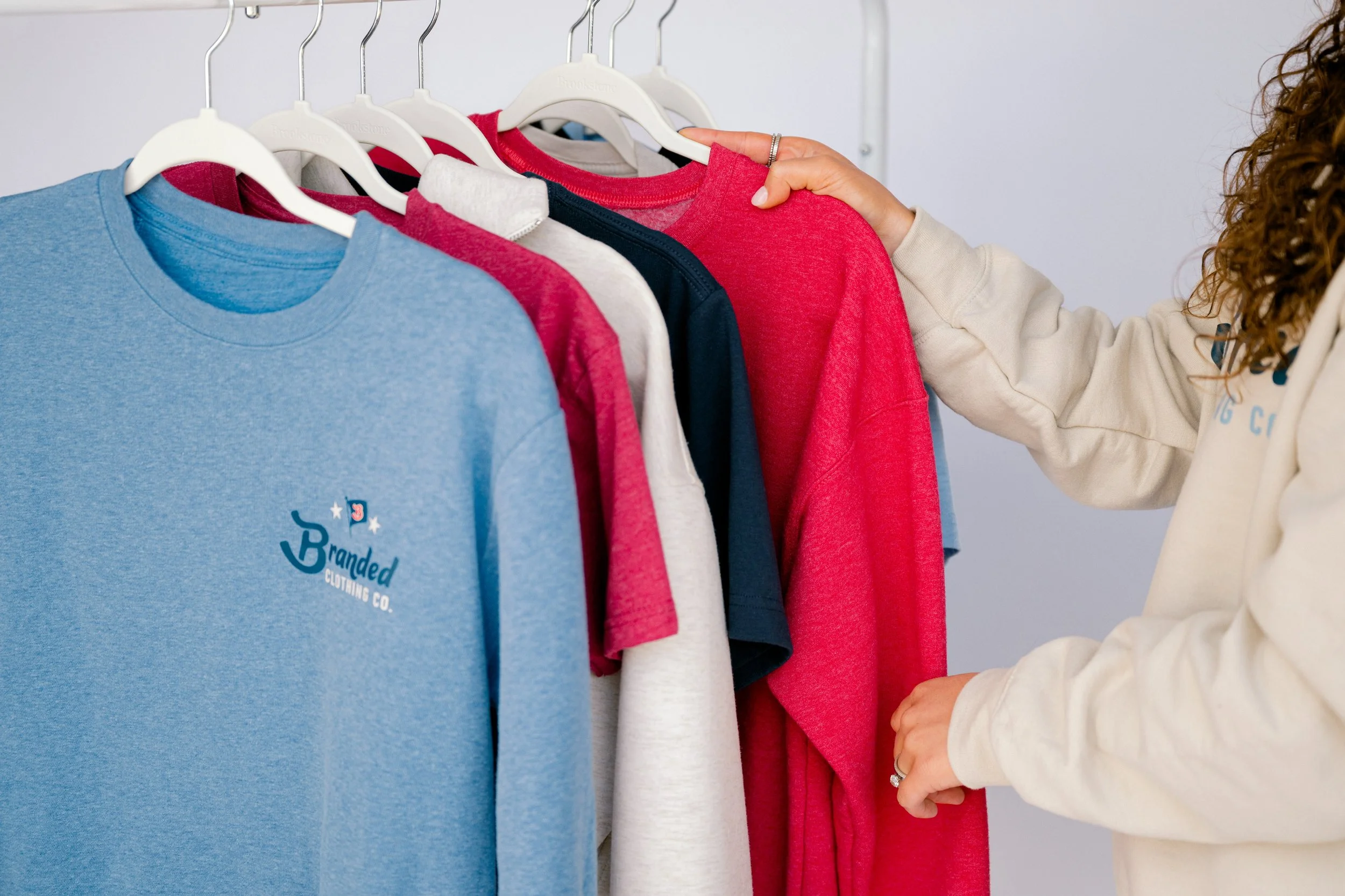 Person selecting a red sweatshirt from a row of colorful t-shirts hanging on a clothing rack.