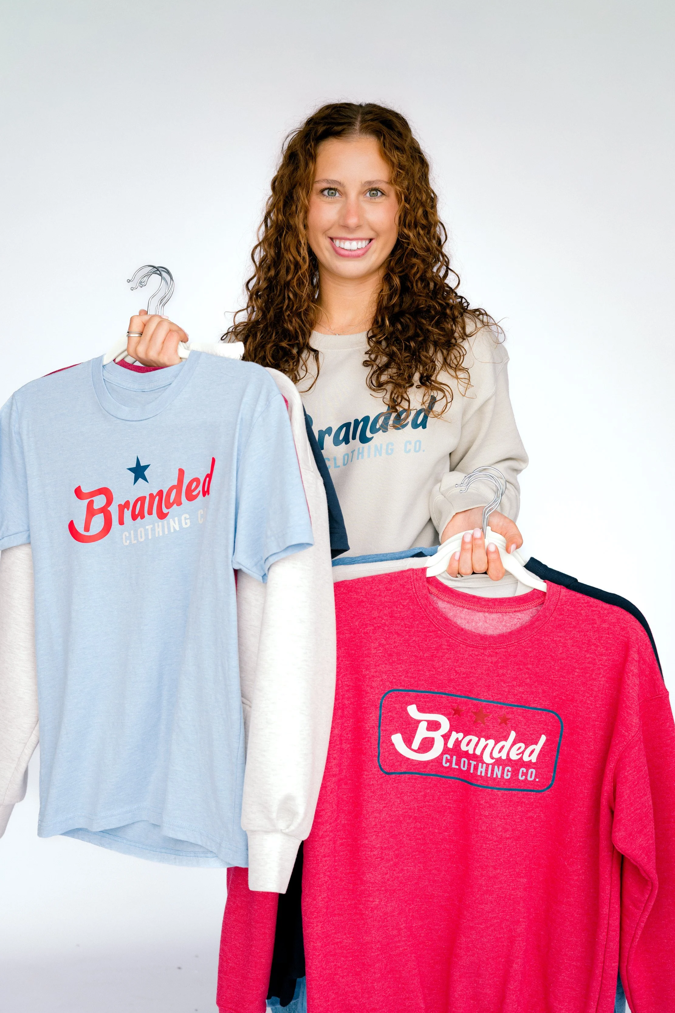 A woman with curly hair holding hangers and wearing a sweatshirt with the text 'Branded Clothing Co.' on it. She is smiling and standing against a plain background.