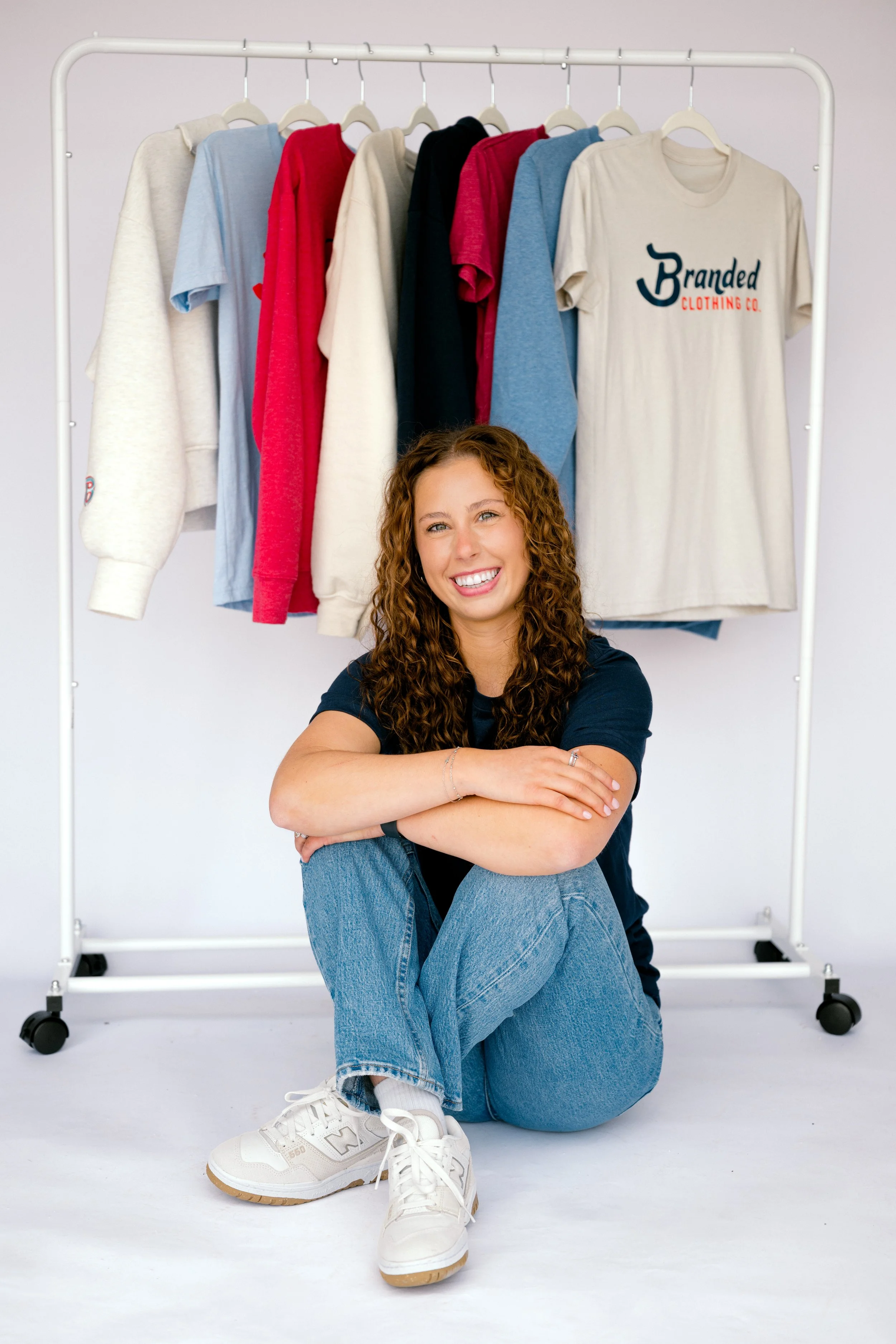 A young woman with curly hair sitting cross-legged in front of a clothing rack with various colored T-shirts and sweatshirts. She is smiling and wearing a dark top, blue jeans, and white sneakers.