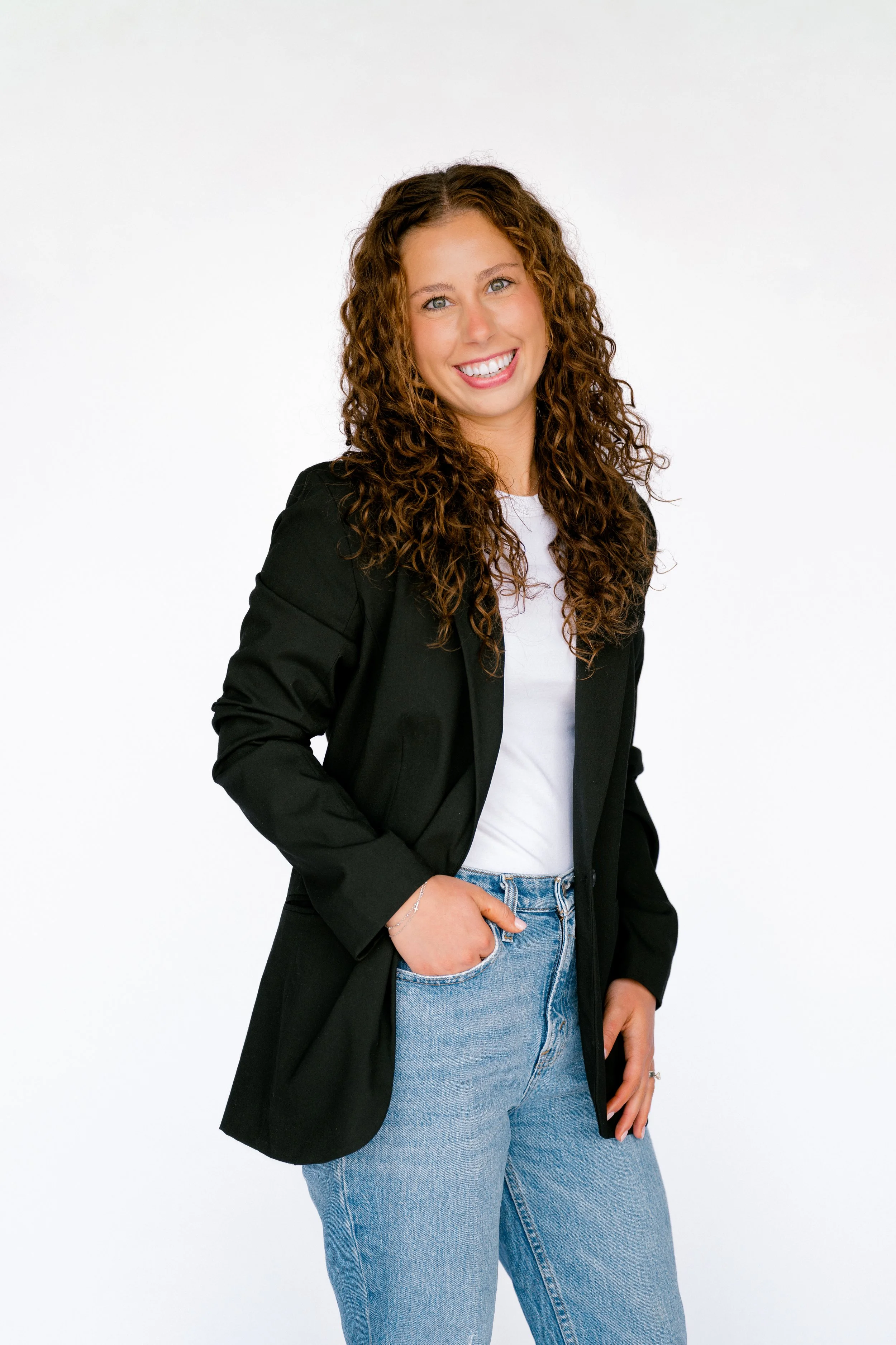 Young woman with curly brown hair smiling, wearing a black blazer, white T-shirt, light blue jeans, and standing against a white background.