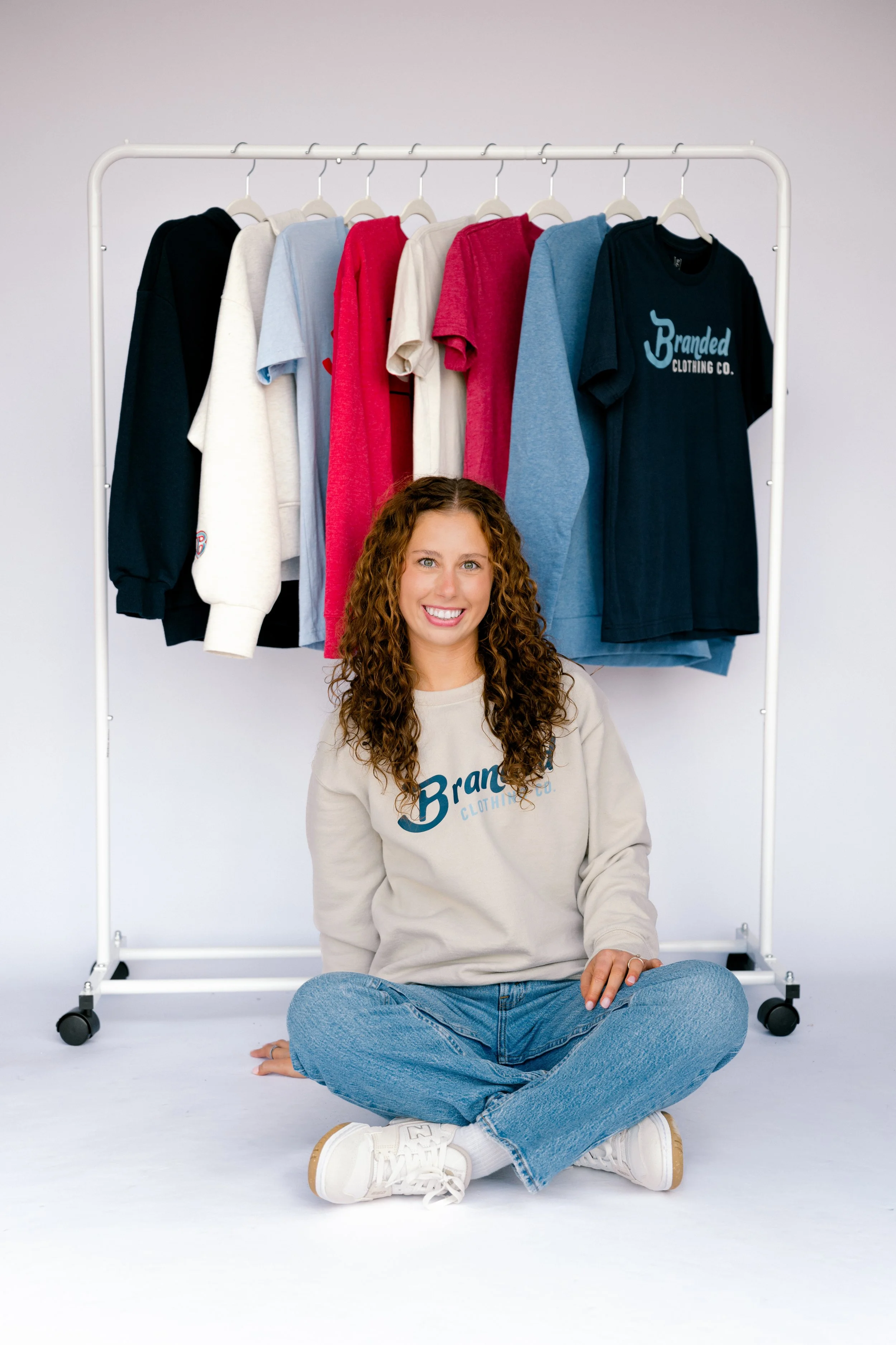 A smiling woman with curly hair sitting cross-legged on the floor in front of a clothing rack with hanging T-shirts in various colors including black, white, red, beige, and blue. The woman is wearing a beige sweatshirt with the logo 'Branded Clothin