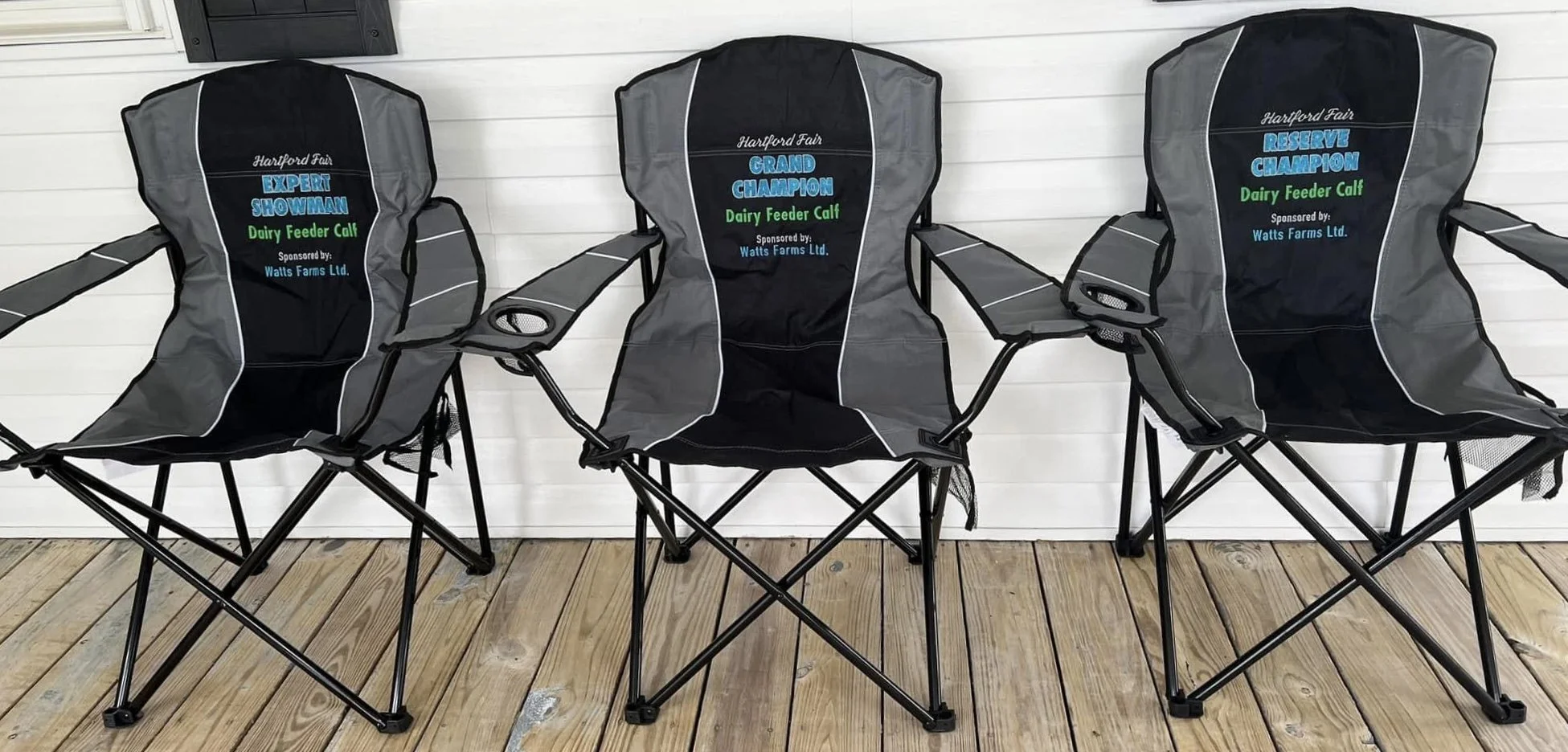 Three black folding chairs with sponsor logos and text for the Hartford Fair, placed on a wooden porch in front of a white wall.