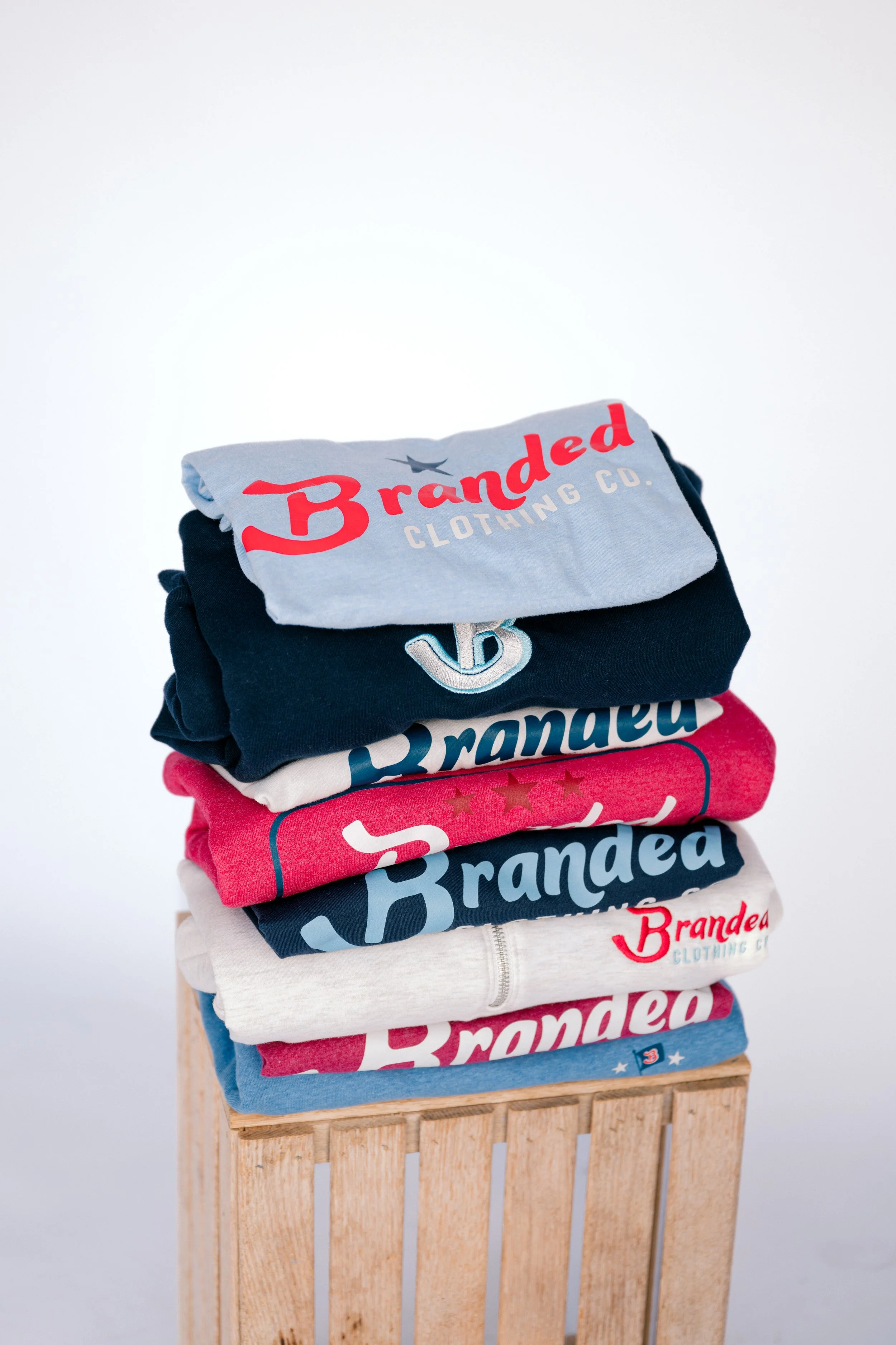 Stack of folded t-shirts with the 'Branded Clothing Co.' logo in red and blue, on a wooden stand against a white background.