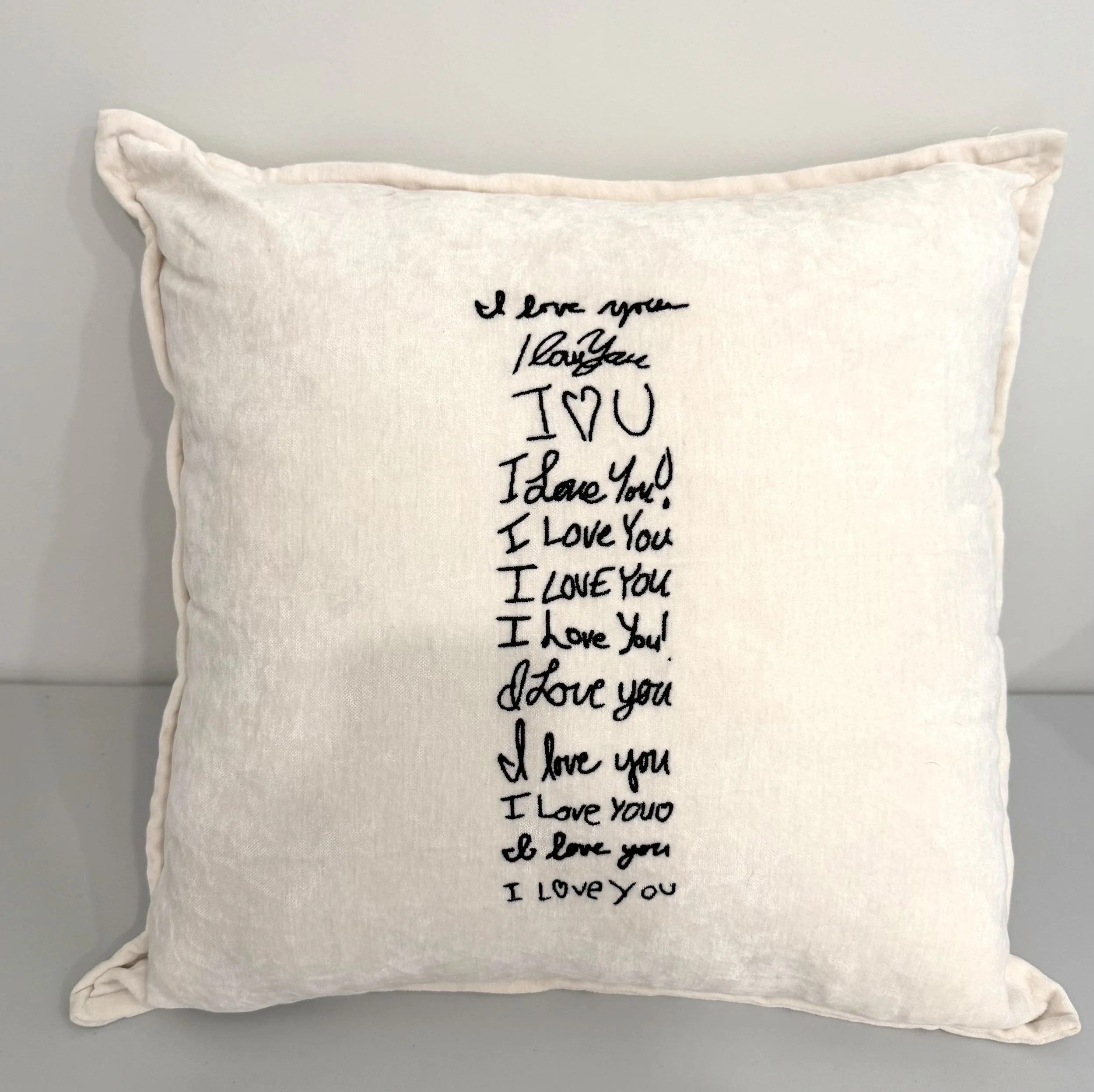 Cream-colored pillow with embroidered message 'I love you' repeatedly in black cursive script, against a light-colored background.