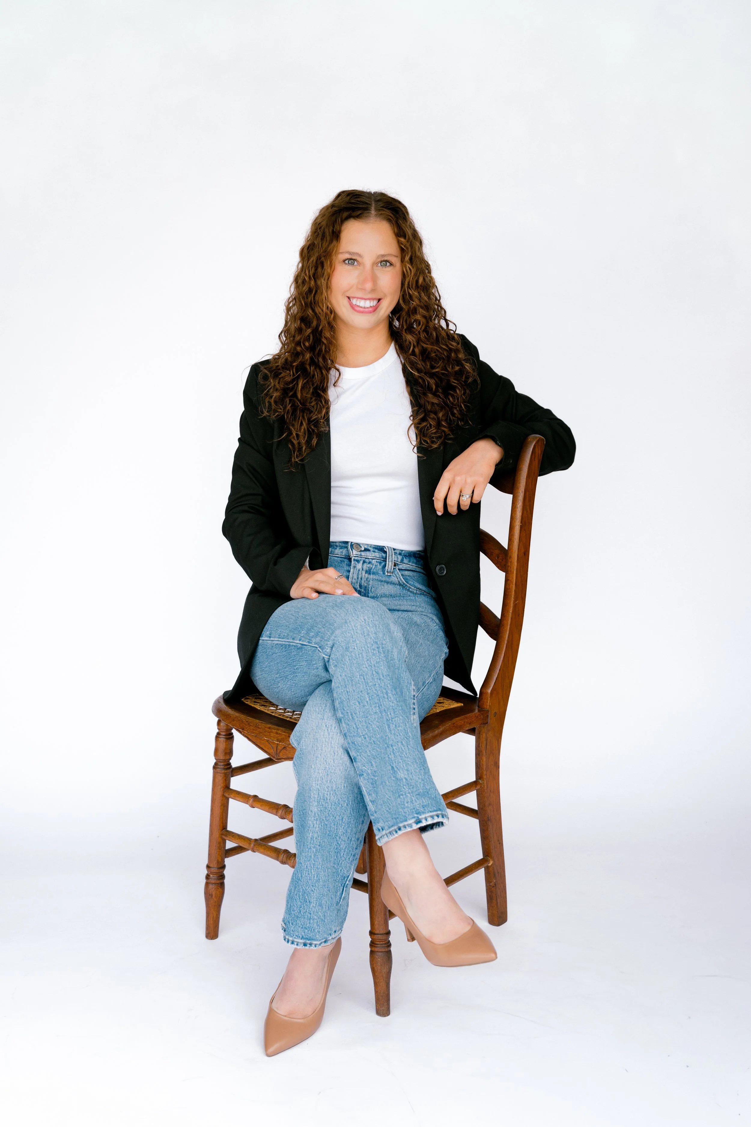A woman with long curly hair wearing a black blazer, white t-shirt, blue jeans, and beige high heels sitting on a wooden chair against a plain white background.