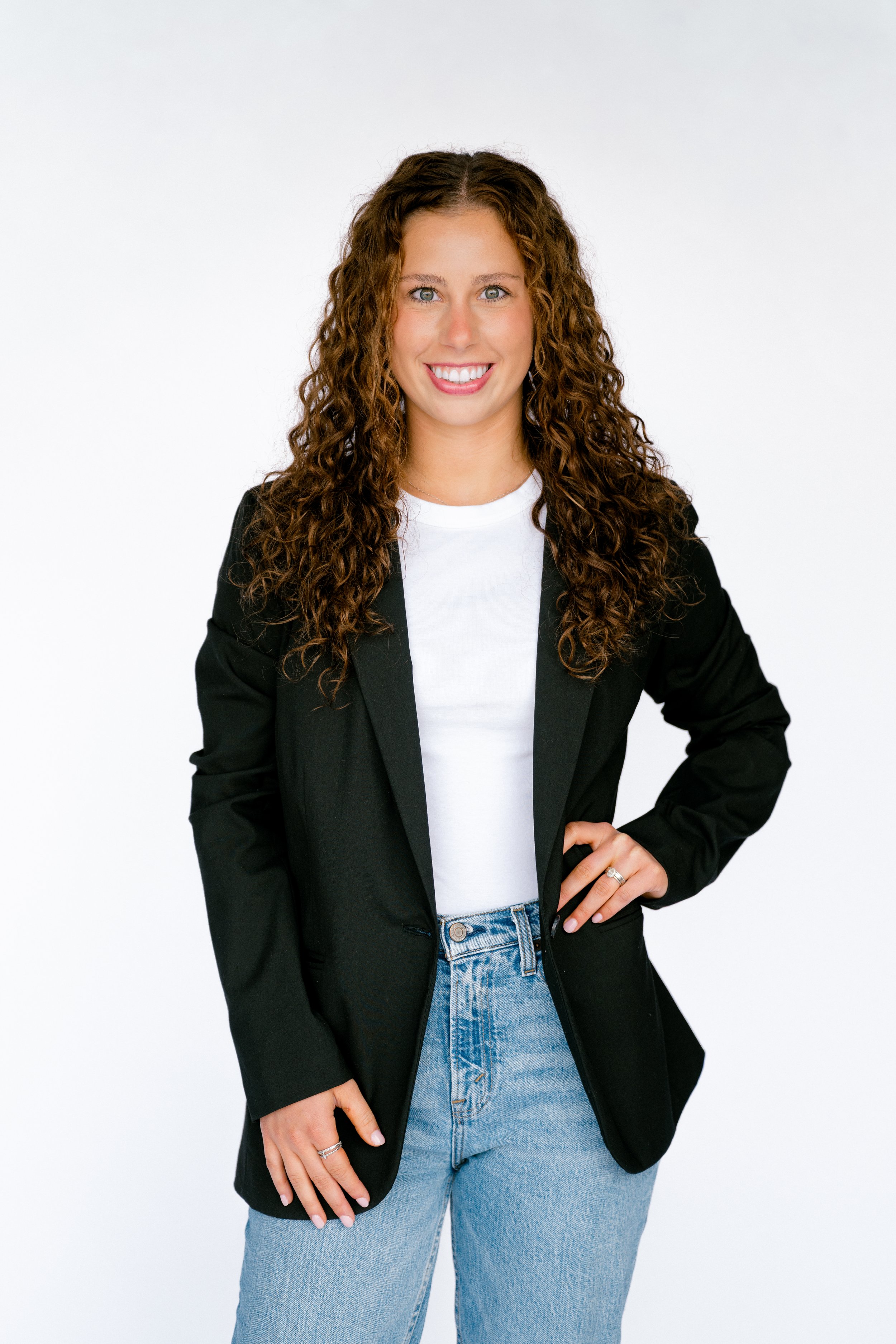 A young woman with curly brown hair, blue eyes, and a bright smile wearing a black blazer, white t-shirt, and light blue jeans, standing against a plain white background.