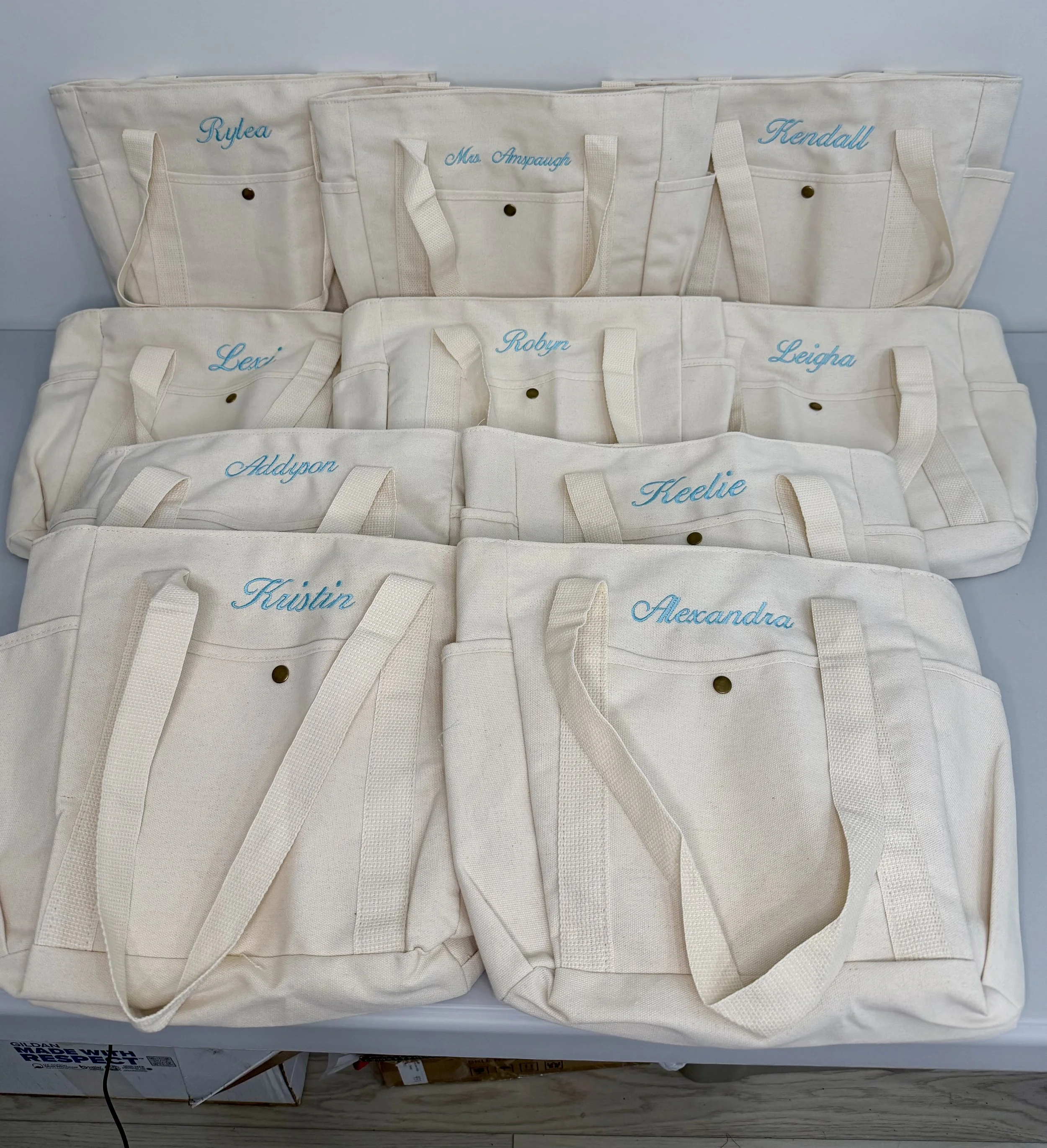 Multiple white canvas tote bags with embroidered blue names, including Rylea, Maddie W, Max Anspaugh, Kendall, Lexi, Robyn, Kelsey, Addyson, and Alexandria, arranged on a flat surface.