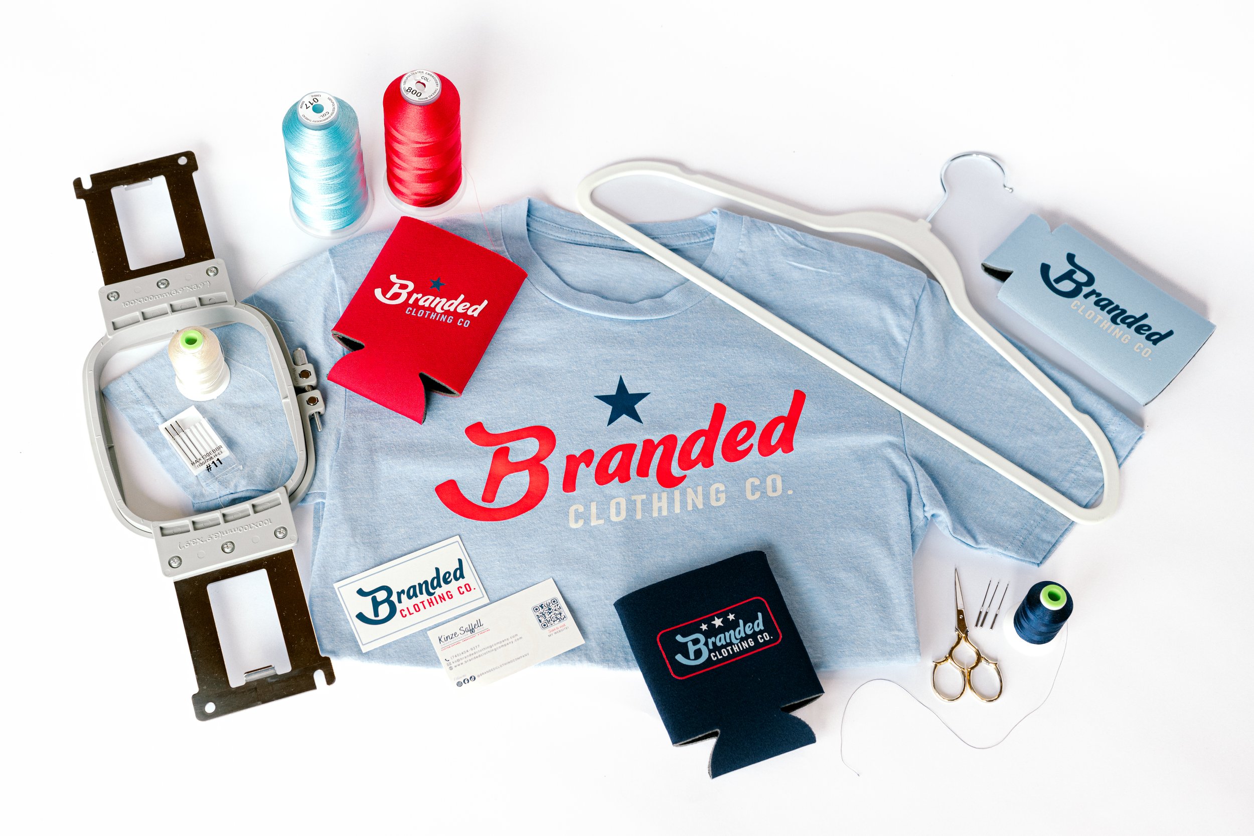 Various sewing and clothing tags, spools of thread in light blue and red, a light blue T-shirt with a branded logo, a plastic hanger, scissors, a needle, and a small black box labeled Branded Clothing Co. on a white background.