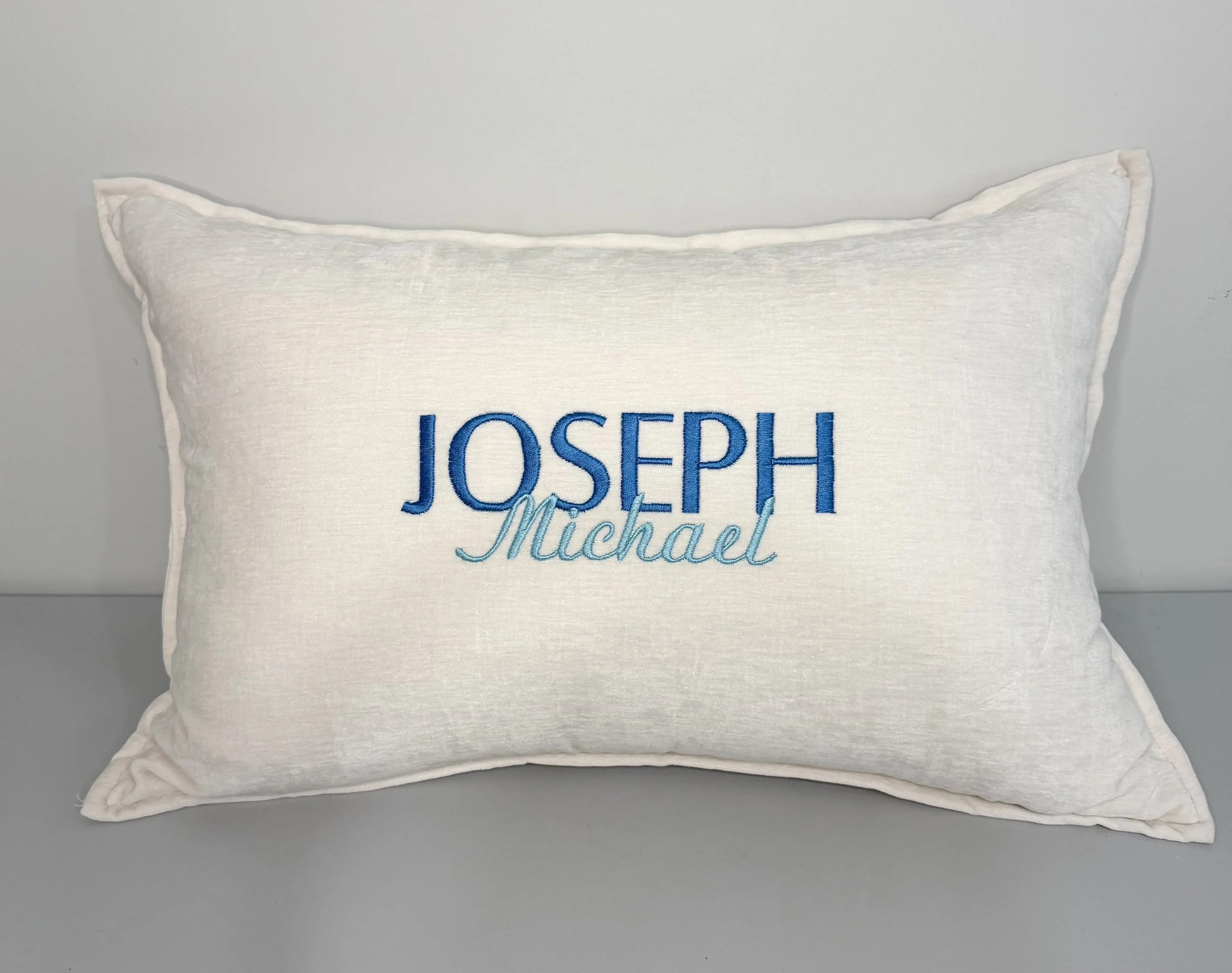 A white pillow with embroidered text 'Joseph Michael' in blue and light blue, placed on a plain light gray surface.