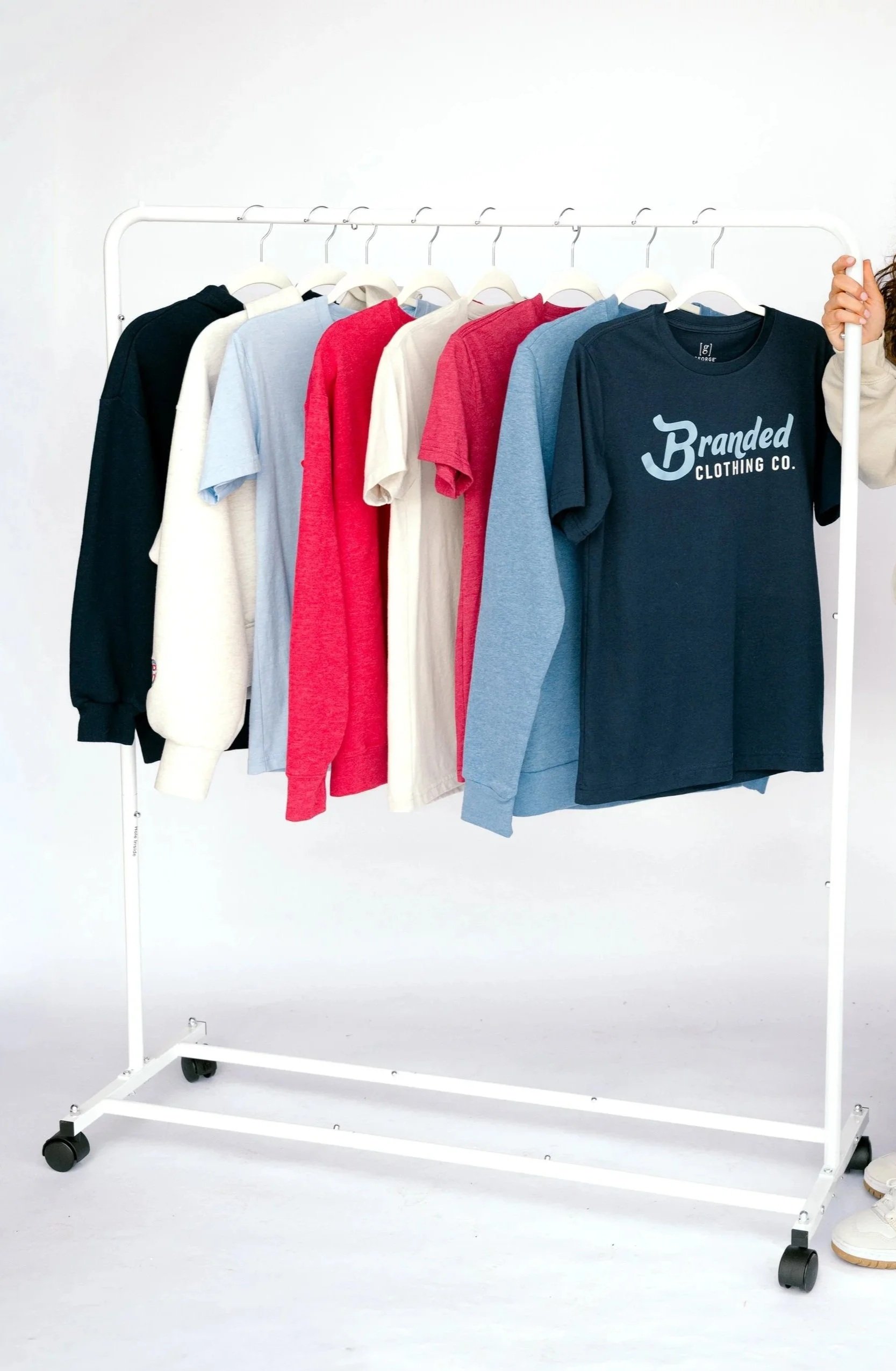 Clothing rack with various T-shirts in black, white, red, cream, light blue, and navy hanging on hangers against a plain white background.