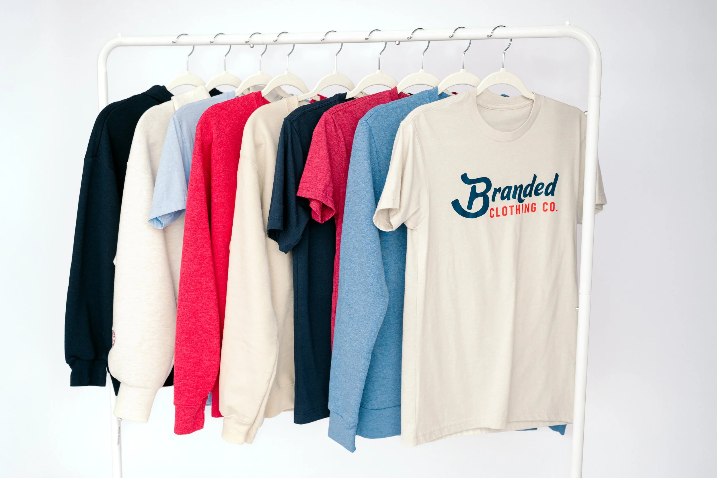 A white clothing rack holding nine t-shirts in various colors including black, white, red, navy, blue, and beige, with one beige t-shirt displaying the logo "Branded Clothing Co."