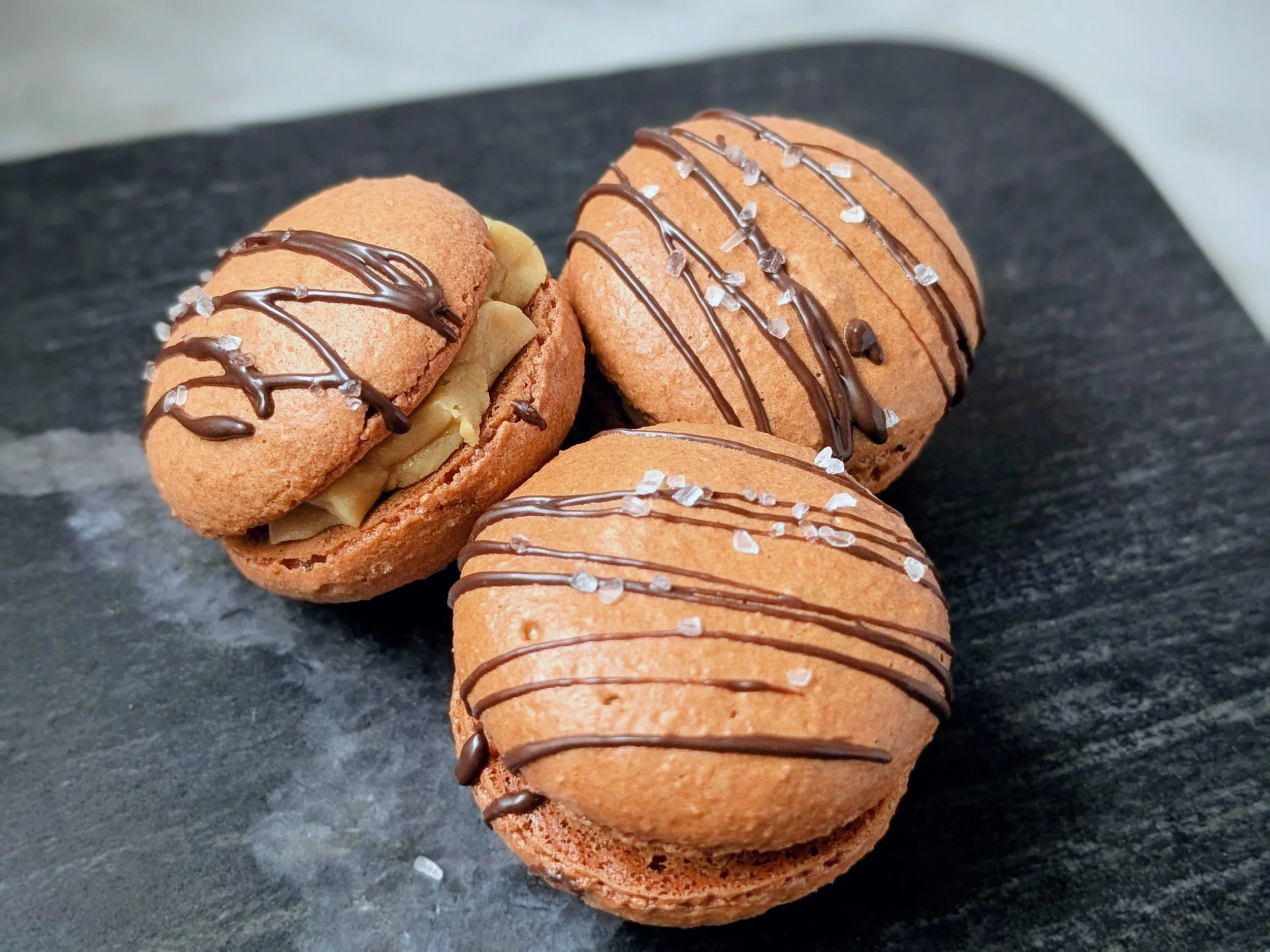 Three macaron cookies with chocolate drizzle and salt flakes on a black slate surface.