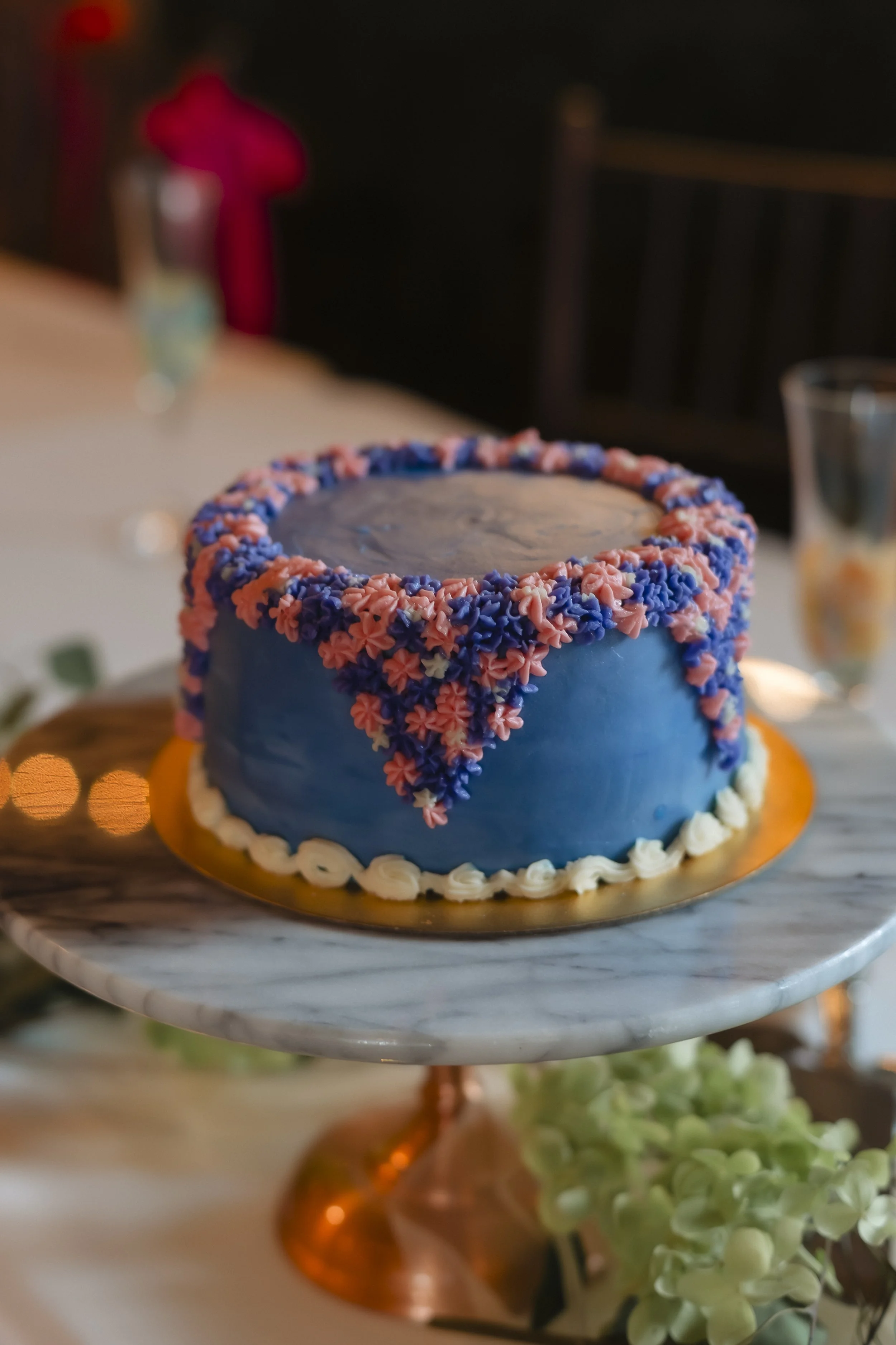 A round birthday cake decorated with pink and purple frosting flowers and blue icing on a marble cake stand.