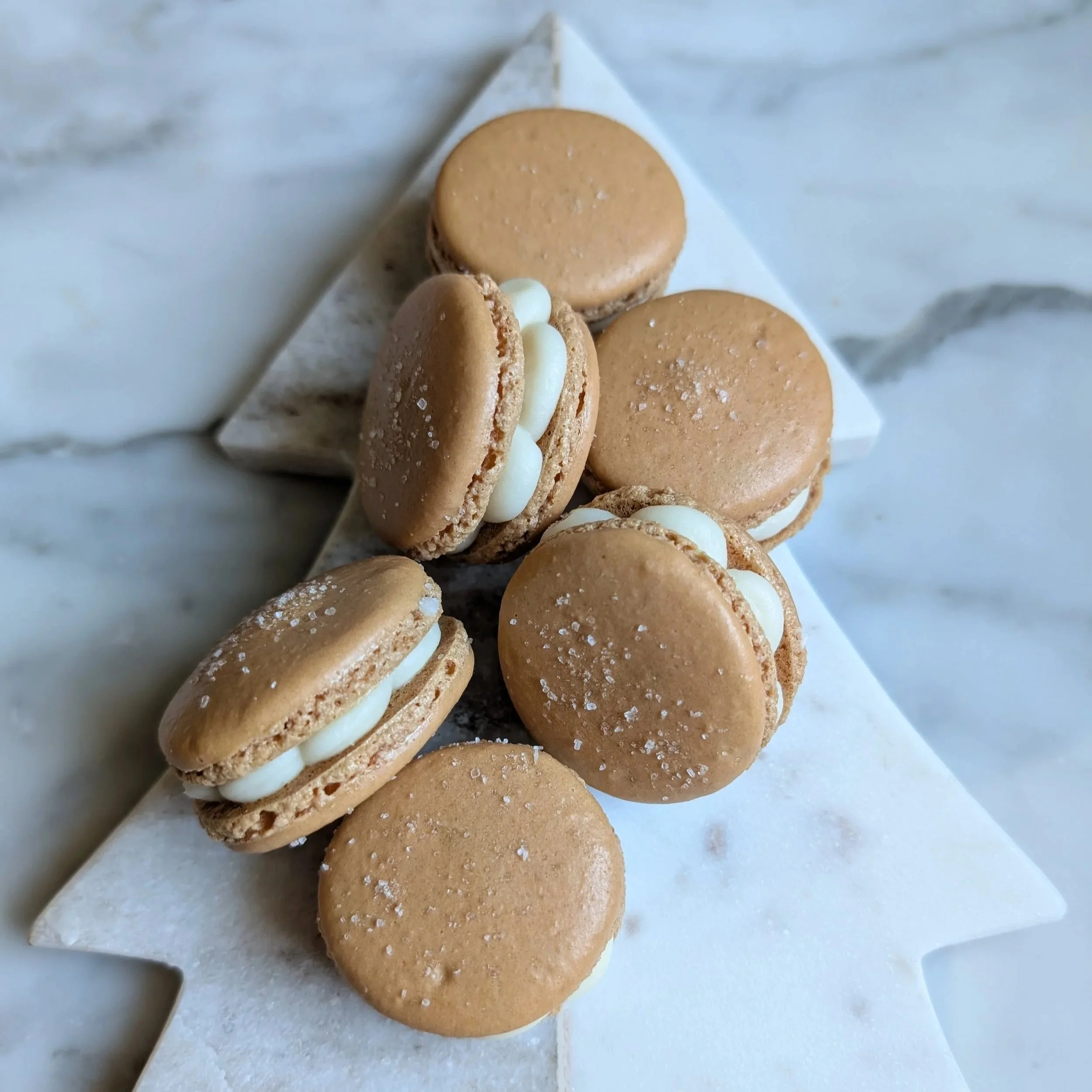 A close-up of several caramel-colored macarons with white filling, arranged on a white marble surface.