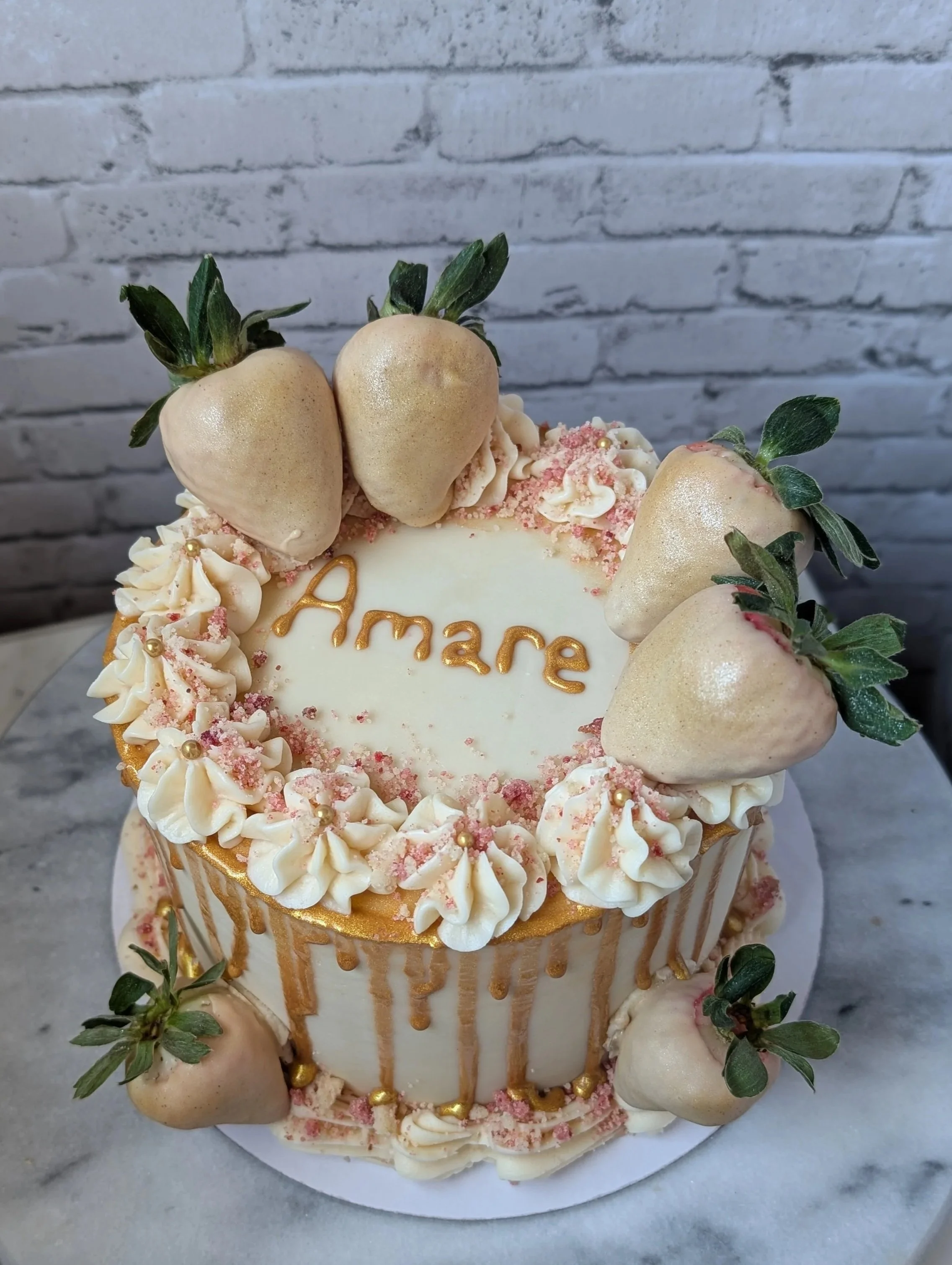 A decorated cake with strawberries, gold accents, and a white and pink frosting border, with the word 'Amare' written on top.