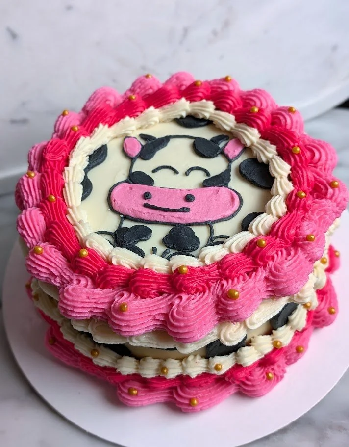 A round two-layer cake decorated with pink and white buttercream frosting, with a cow illustration on top made of icing, featuring black spots and a pink snout, adorned with small golden edible beads.
