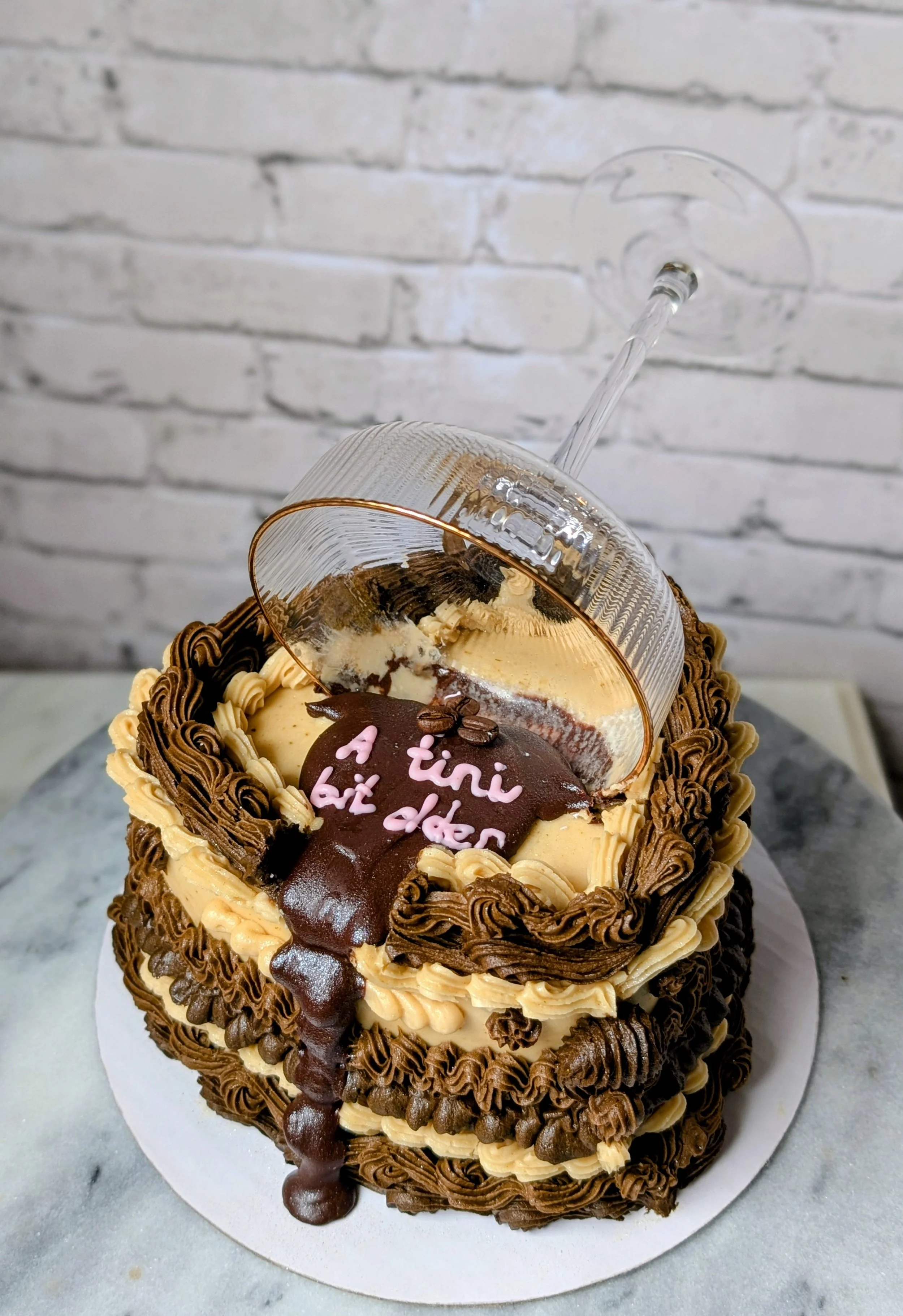 A heart-shaped chocolate and vanilla layered cake with decorative swirls of frosting, topped with a chocolate plaque with the message 'a tiny bit d**ed' in pink and a small piping of frosting, and a transparent plastic glass upside down on top.