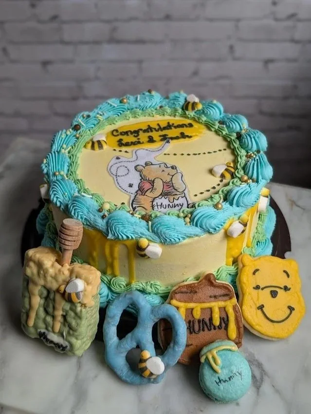 A round cake with a honey and bee theme, decorated with blue, green, and yellow frosting. The top has an edible image of Winnie the Pooh and a bee, with the message 'Congratulations Laci & Jaci.' Surrounding the cake are honey-themed decorations including a honey dipper, honeycomb, a honey jar labeled 'Hunny,' a blue ring, a blue and yellow ball labeled 'Hunny,' and a smiling bear face.