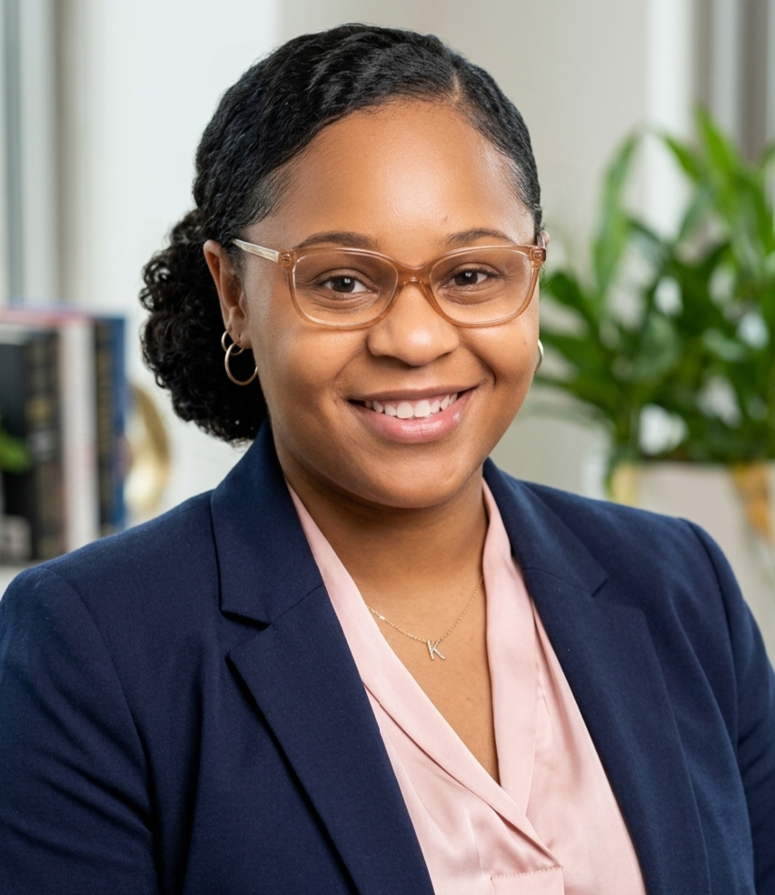 A woman with glasses and earrings, wearing a navy blazer and pink blouse, smiling at the camera, with a background of a bookshelf and a plant.