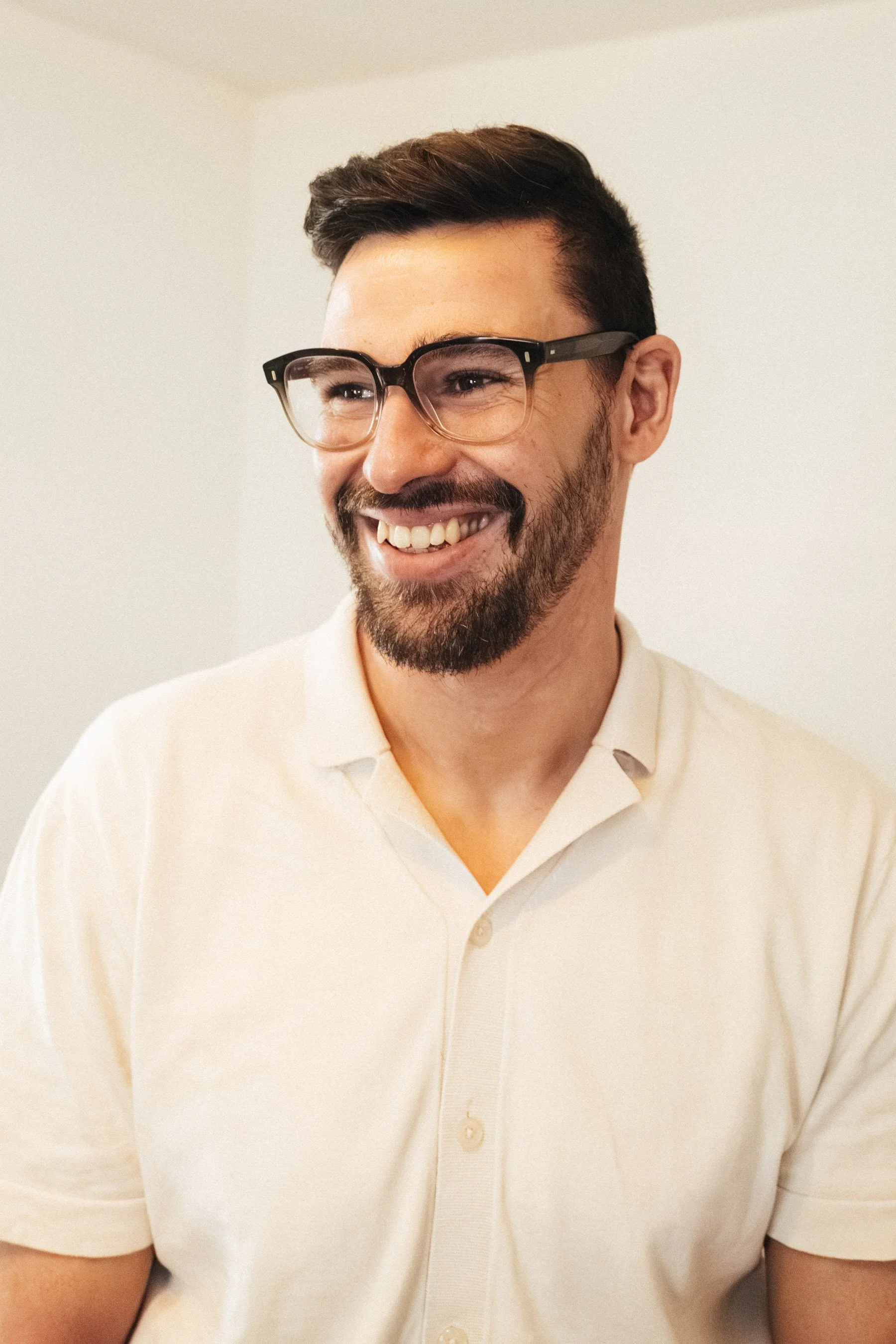 Ein lachender Mann mit Brille und dunklem Bart trägt ein weißes Hemd in einem Raum mit neutraler Wandfarbe.