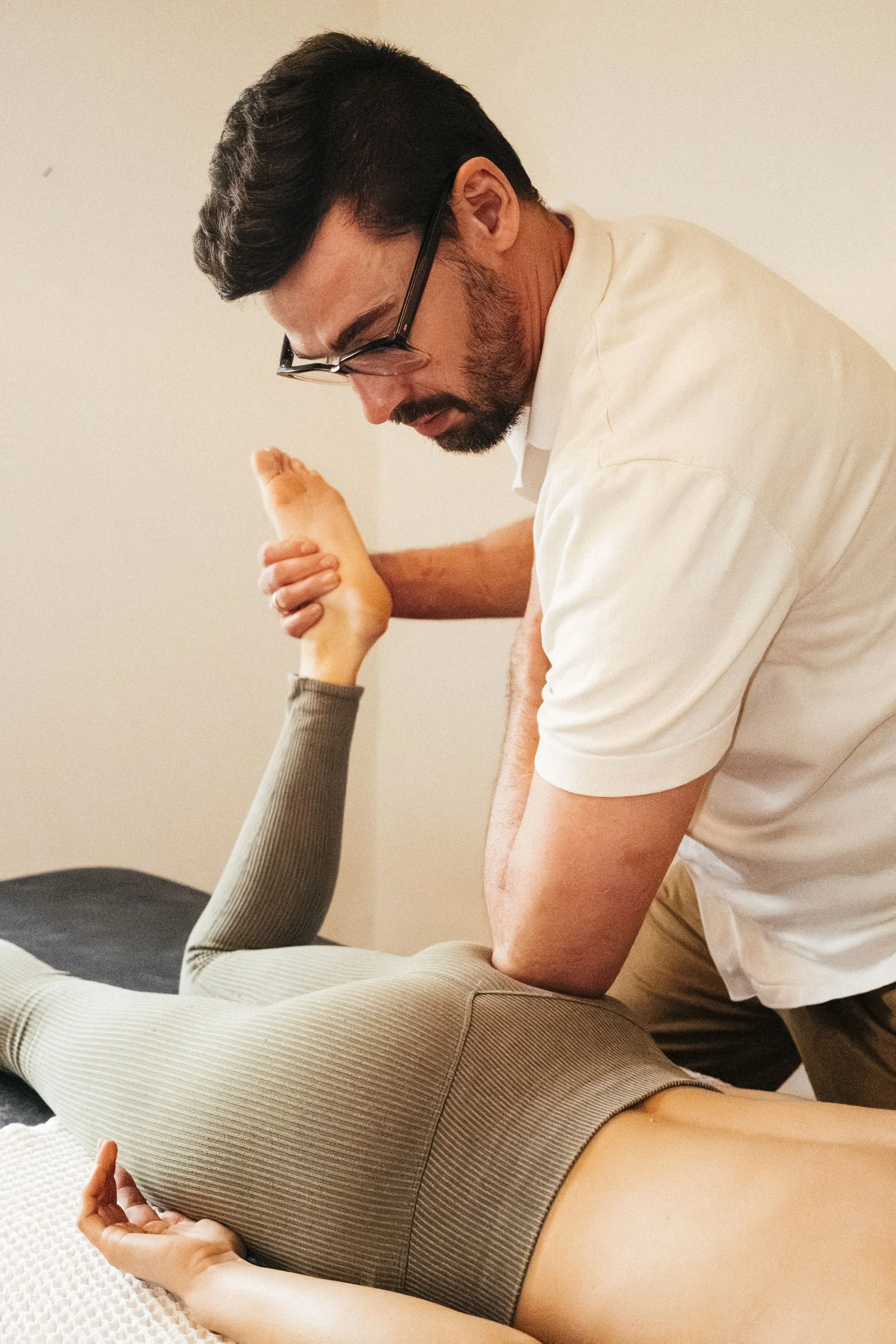 Ein Mann mit Brille führt eine physiotherapeutische Übung bei einer liegenden Person durch.
