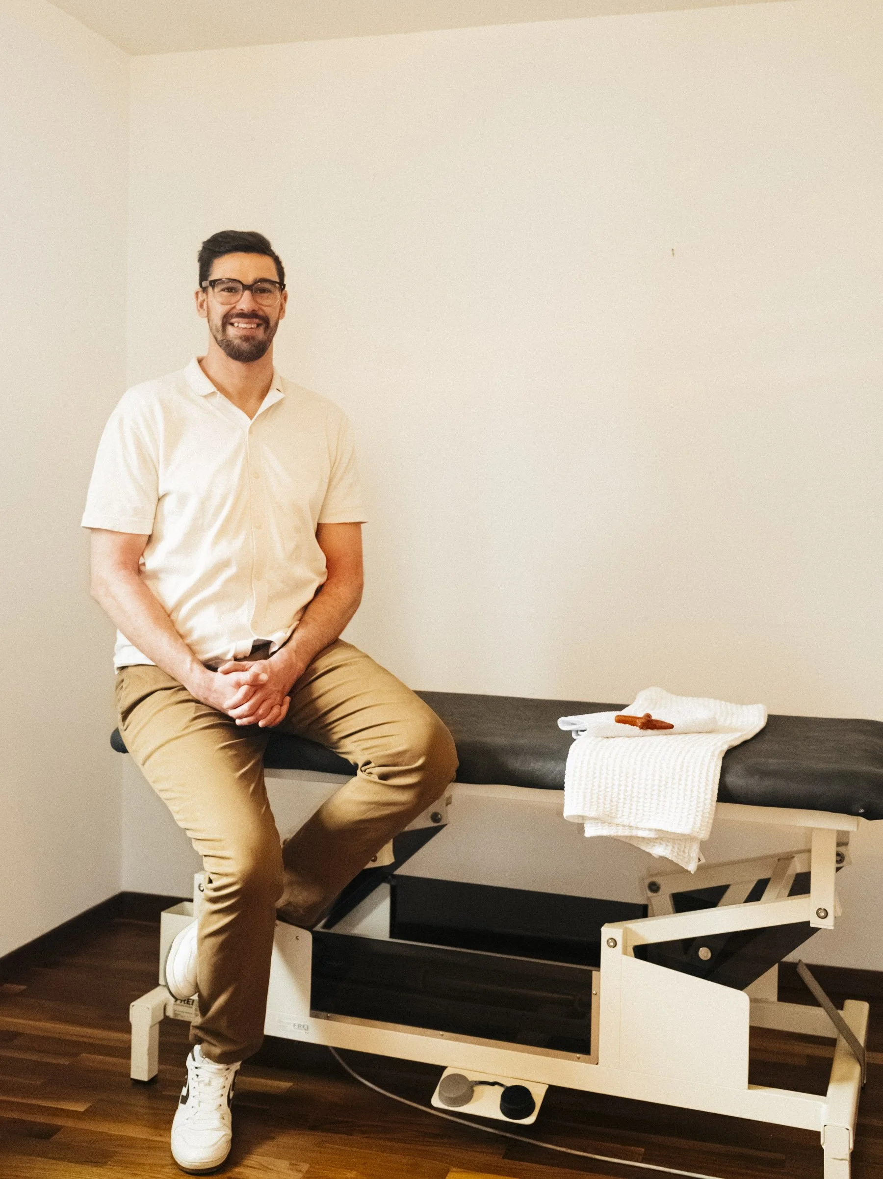 Ein Mann sitzt lächelnd auf einem medizinischen Behandlungstisch in einem Raum, der auf eine chiropraktische oder physiotherapeutische Behandlung hinweisen könnte, mit einem weißen Handtuch und einem kleinen Gegenstand darauf.