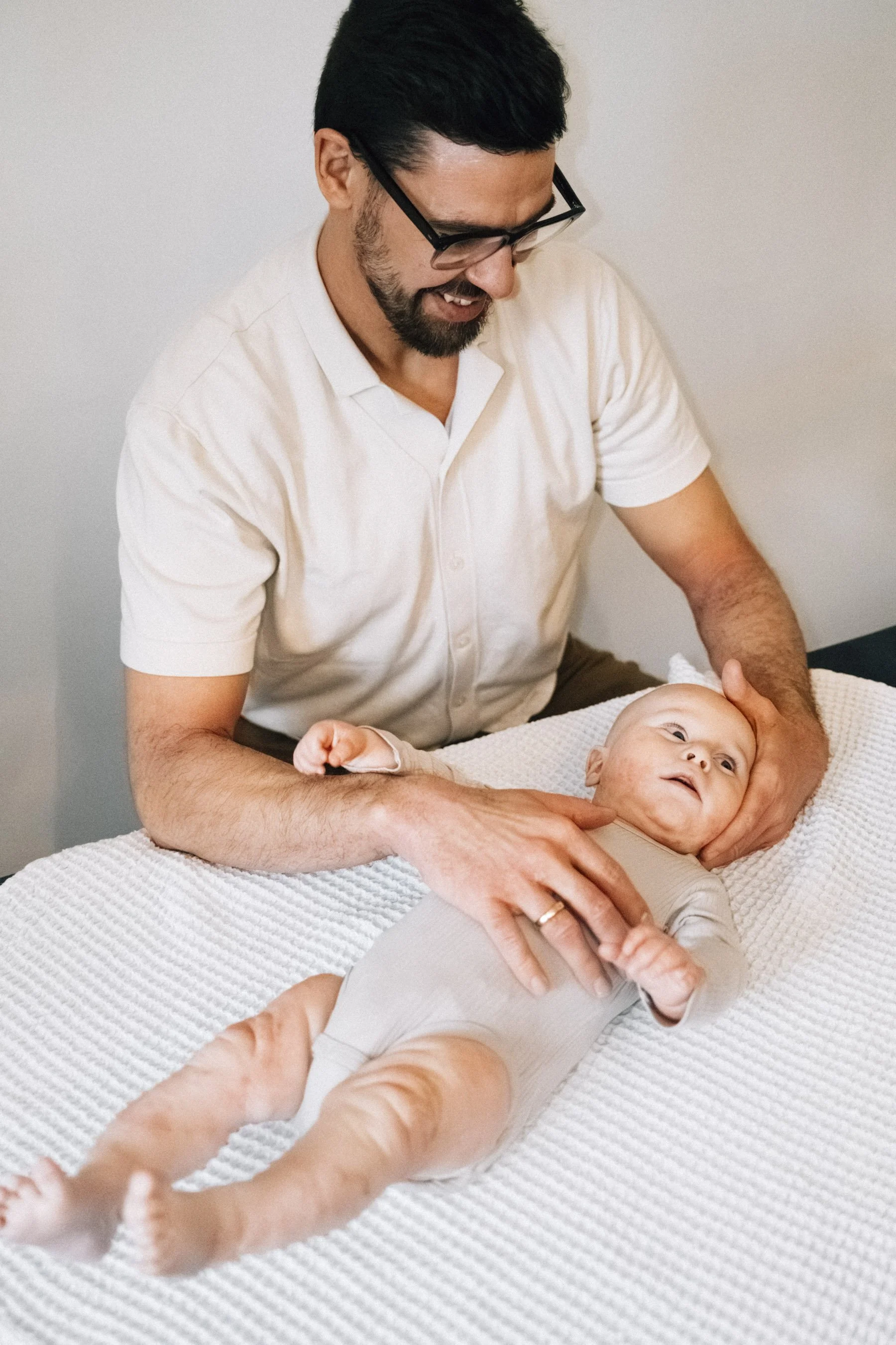 Ein Mann, vermutlich ein Vater, hält ein Baby auf einem Wickeltisch. Der Mann trägt eine weiße Hemd, trägt eine Brille und lächelt. Das Baby liegt auf dem Rücken, trägt einen beigen Body und schaut nach oben.