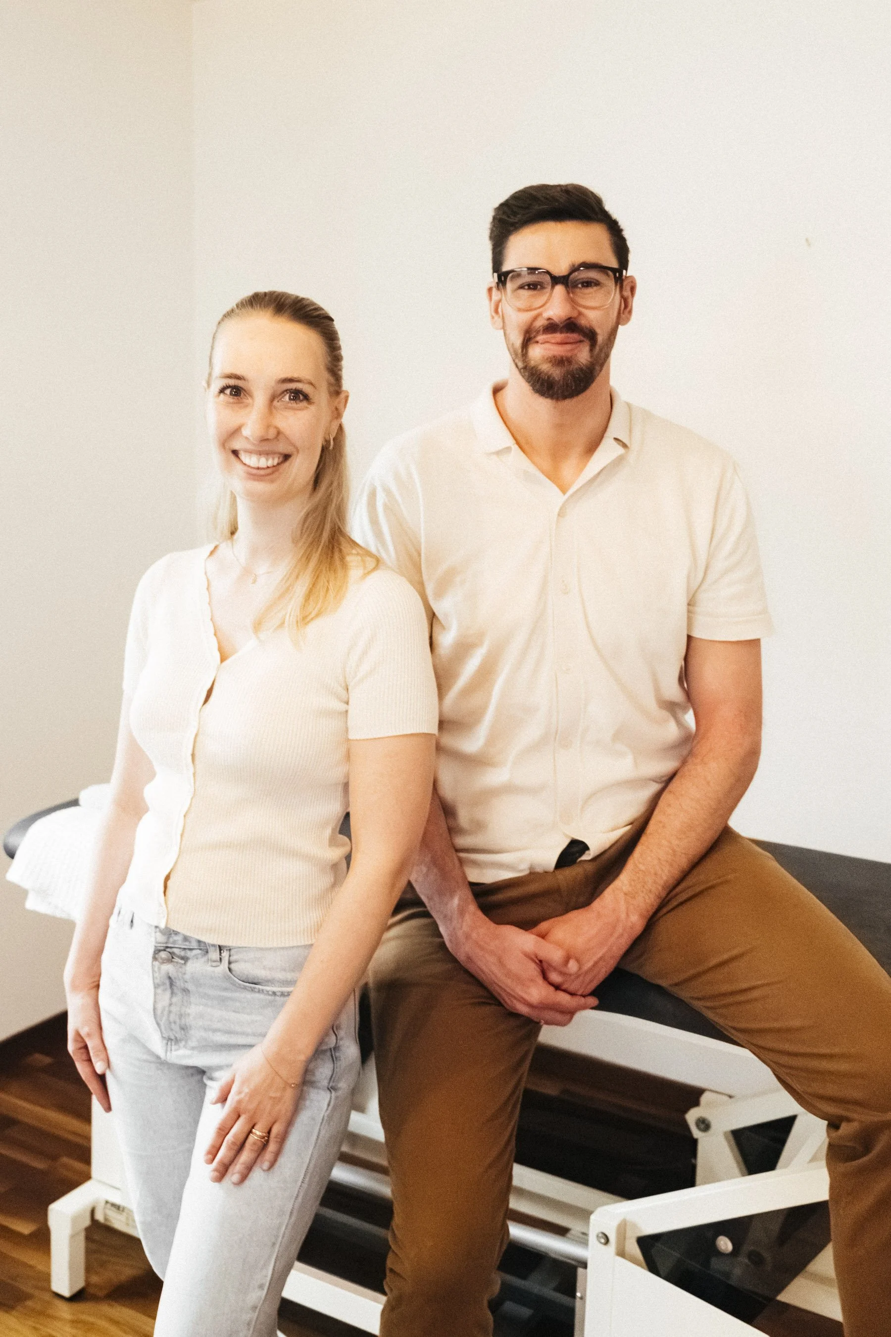 Zwei Menschen in einem medizinischen Raum, eine Frau und ein Mann, posieren für ein Foto, während sie neben einem Untersuchungstisch sitzen.