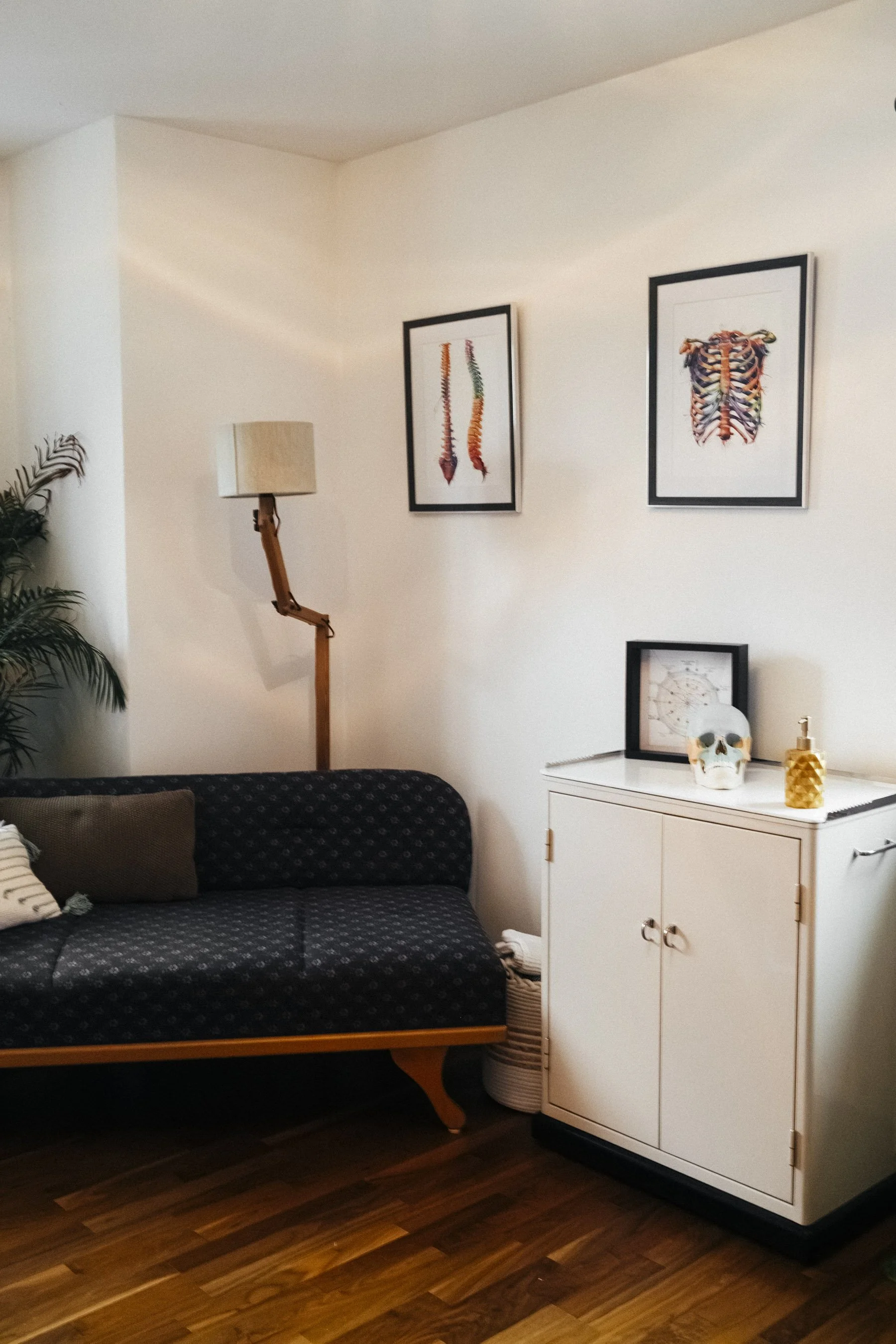 Ein Wohnzimmer mit einer schwarzen Couch, einer weißen Kommode mit Dekorationen, Wandbildern über der Kommode und Pflanzen, Holzboden.