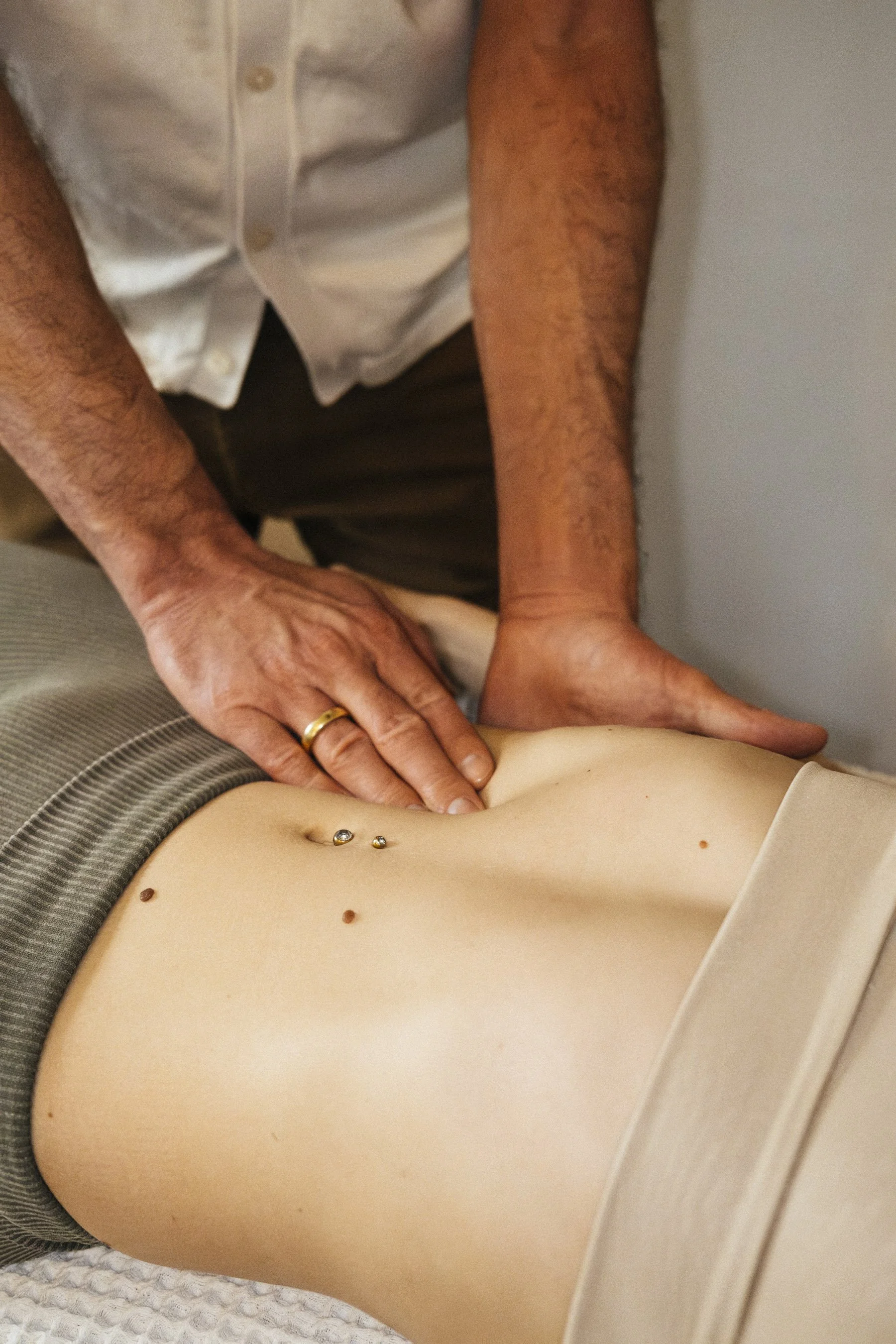 Eine Person macht eine Bauchmassage, während eine andere Person liegt. Die Person, die die Massage durchführt, trägt ein weißes Hemd, während die liegende Person einen grauen Beanie trägt. Es sind Piercings sichtbar auf dem Bauch der liegenden Person.