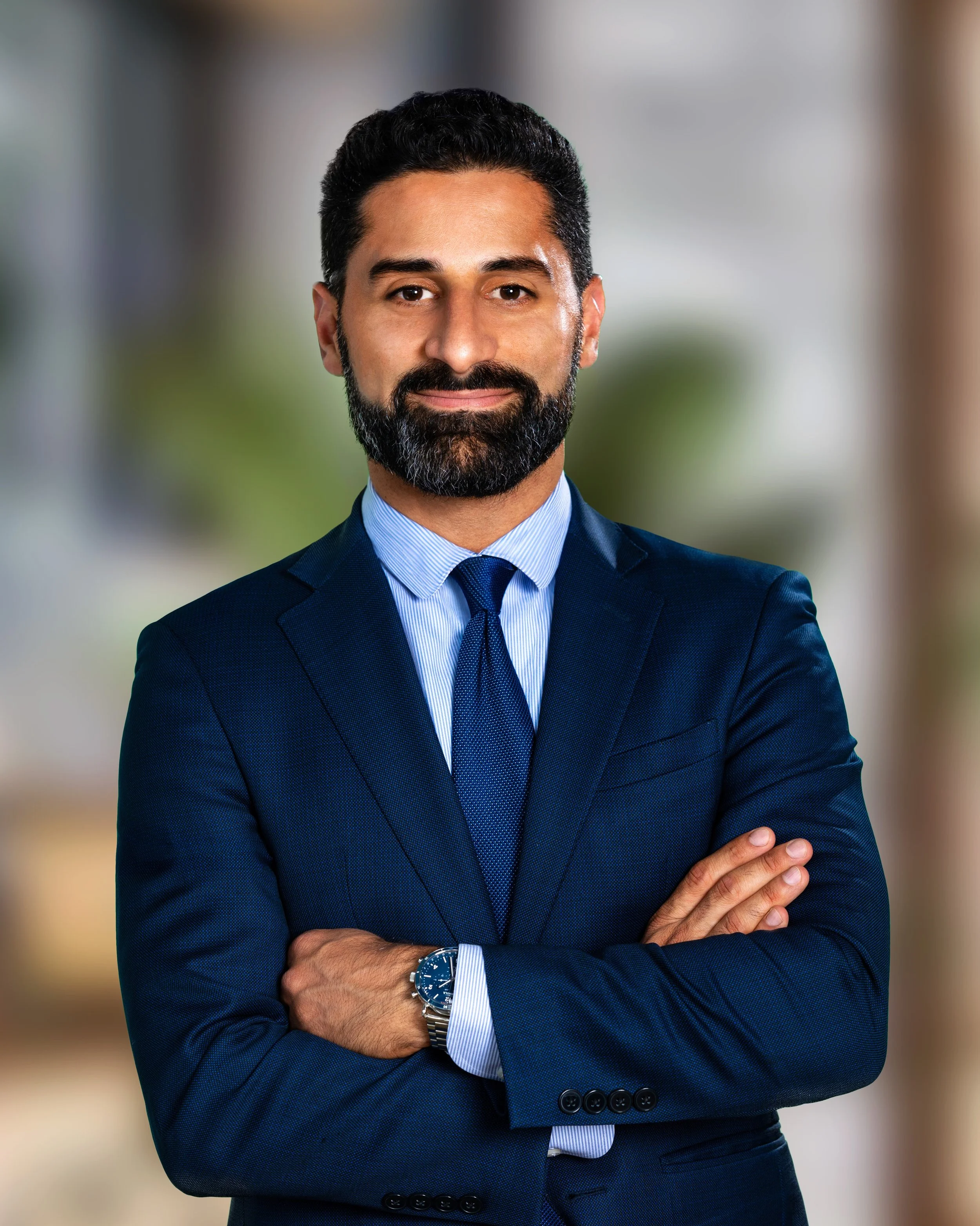 A man with dark hair and a beard wearing a navy blue suit, light blue shirt, and matching tie, posing with his arms crossed in an indoor setting with blurred background.
