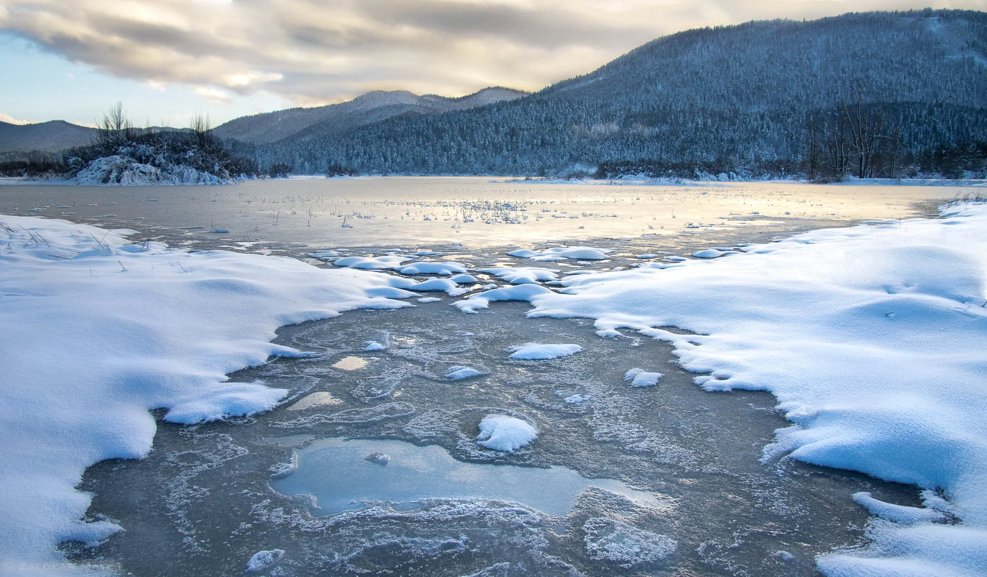 Frozen winter landscape with snow-covered ground and icy river, mountains in the background, cloudy sky.