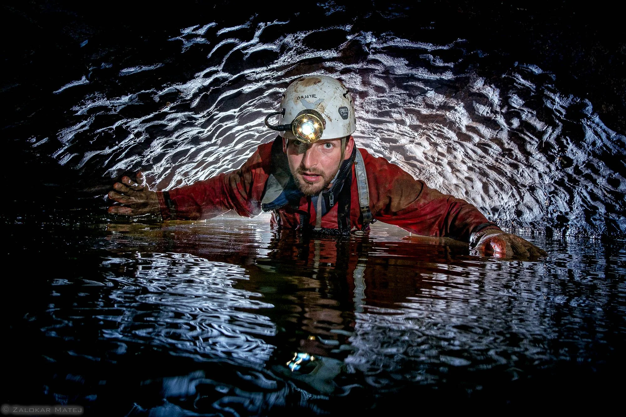A male caving explorer with a headlamp crawling through a dark, wet underground tunnel.