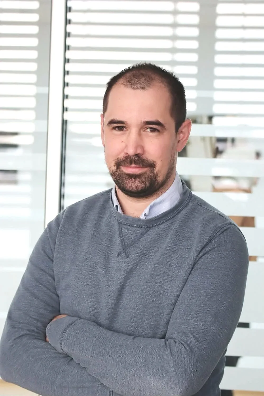 A man with a beard and mustache, wearing a gray sweater and shirt, standing with arms crossed in front of window blinds.