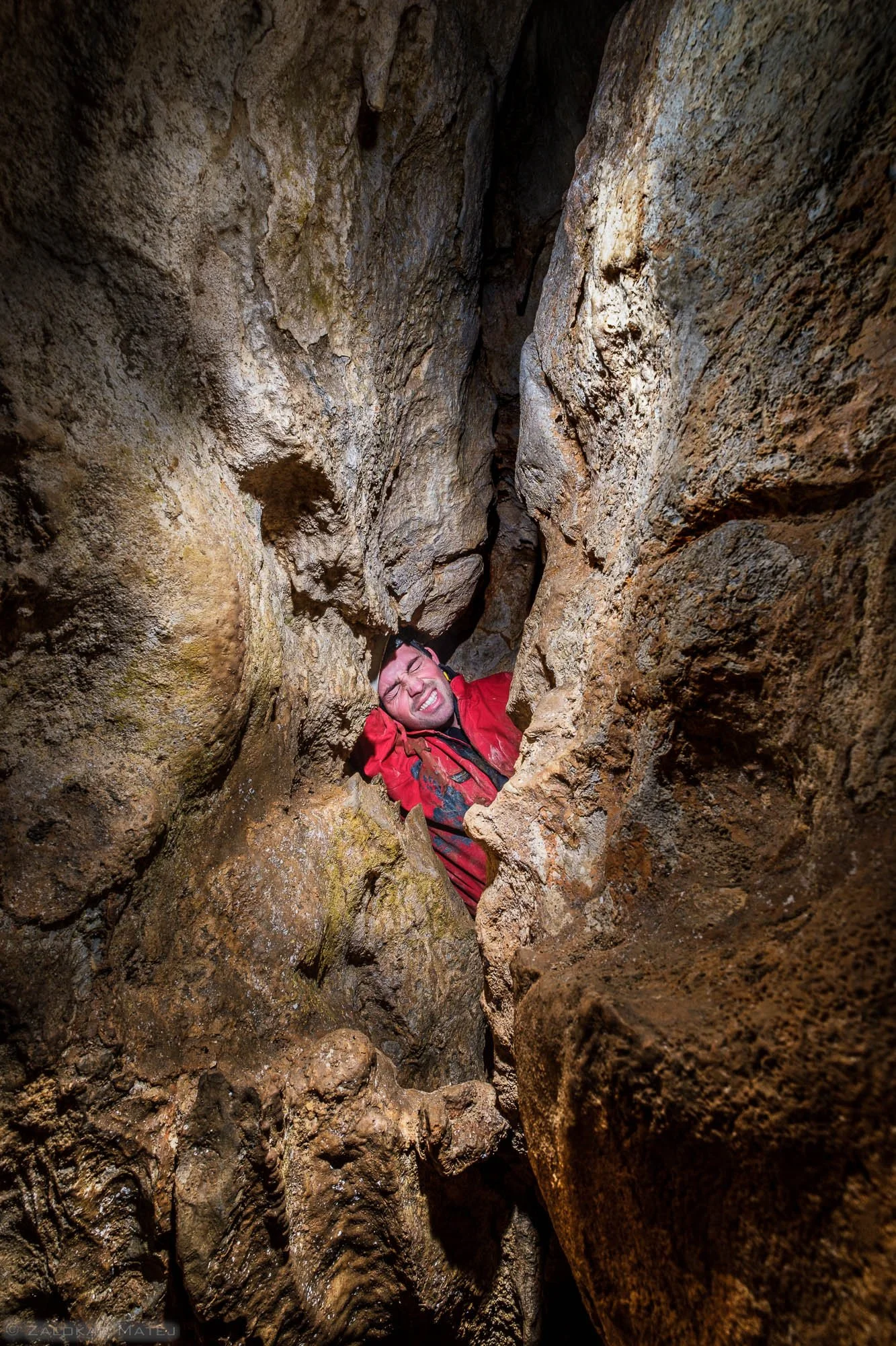 A man in a red jacket is stuck between rocks in a narrow cave passage, with a strained expression on his face.