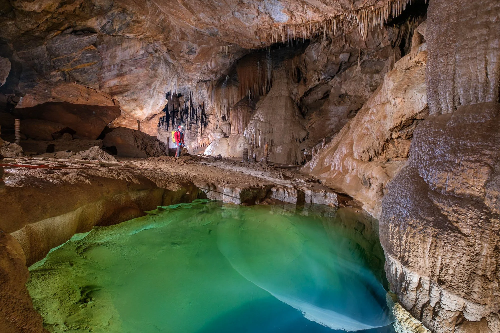 A person in outdoor gear with a helmet inside a large underground cave standing near a small pool of green water, surrounded by stalactites and stalagmites.