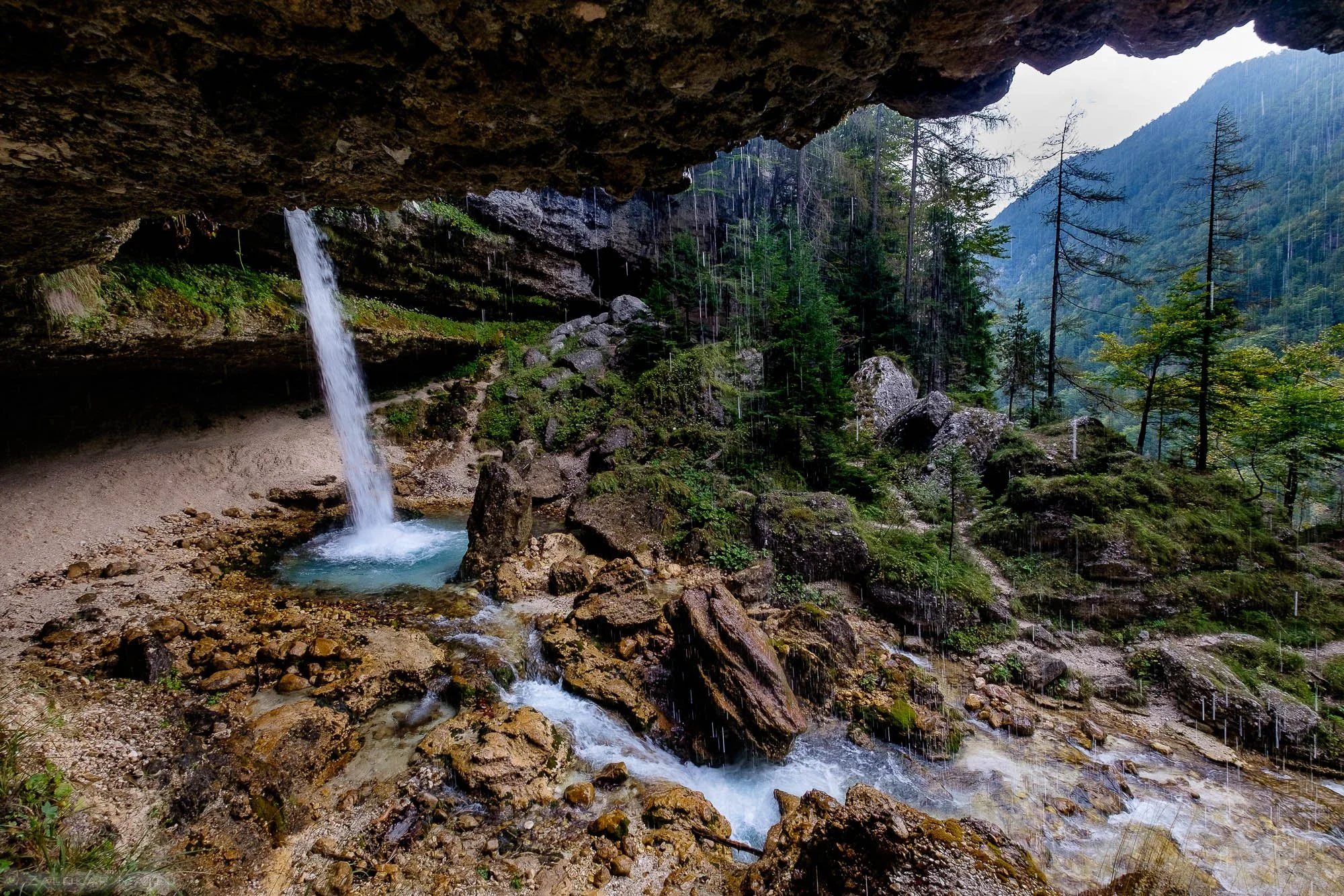 View of a small waterfall cascading under a rocky overhang into a rocky stream in a forested mountainous area with tall trees and mountains in the background during rainfall.