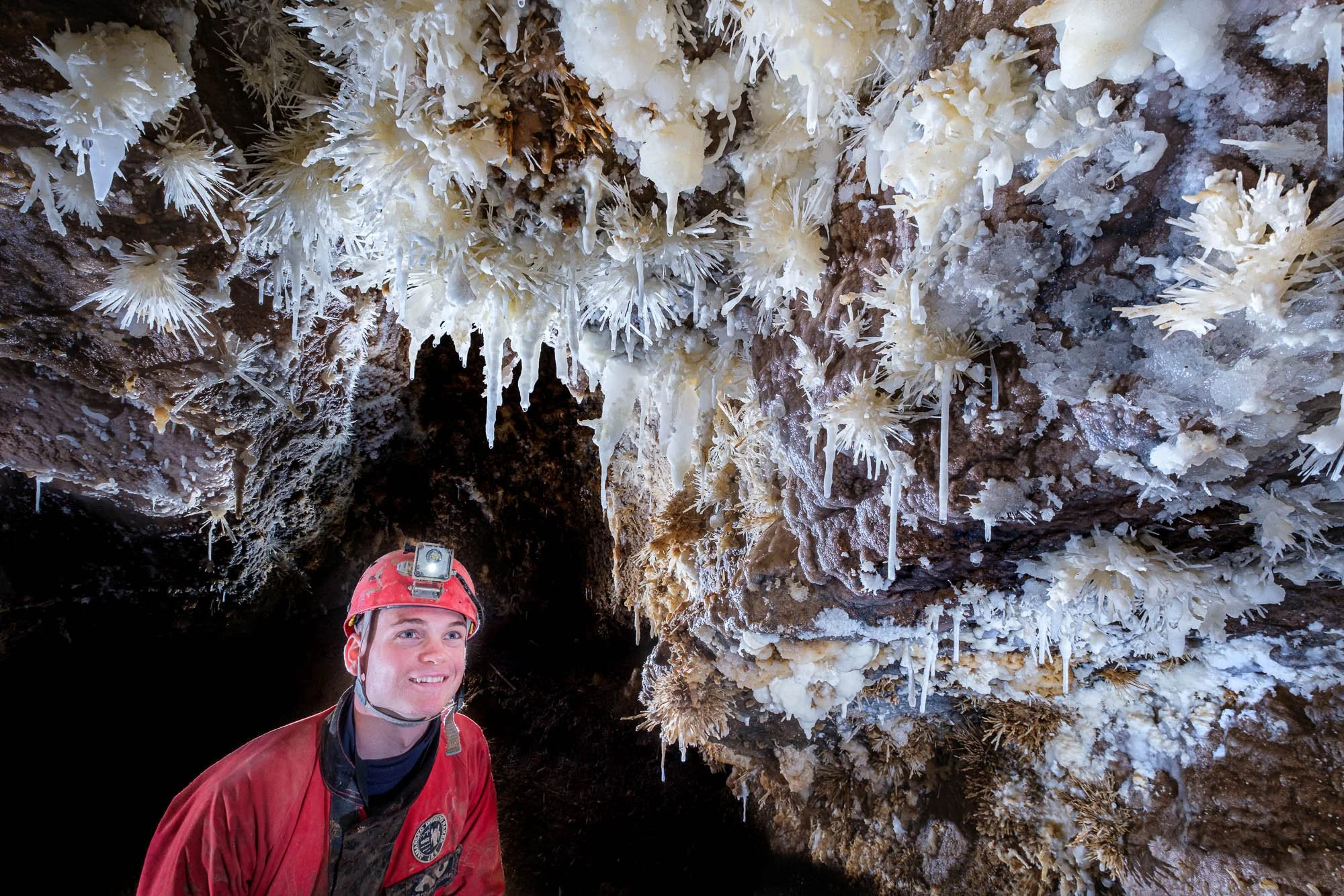 A person in caving gear, wearing a red helmet with a headlamp, inside a cave with white mineral formations on the ceiling.