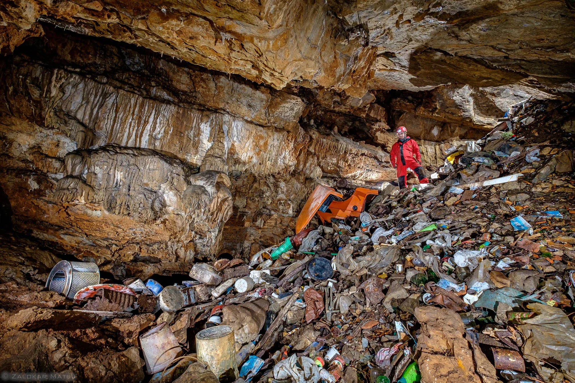 A person in red jacket and helmet standing in a cave filled with a large amount of trash, including plastic bottles and cans, accumulated on the floor.