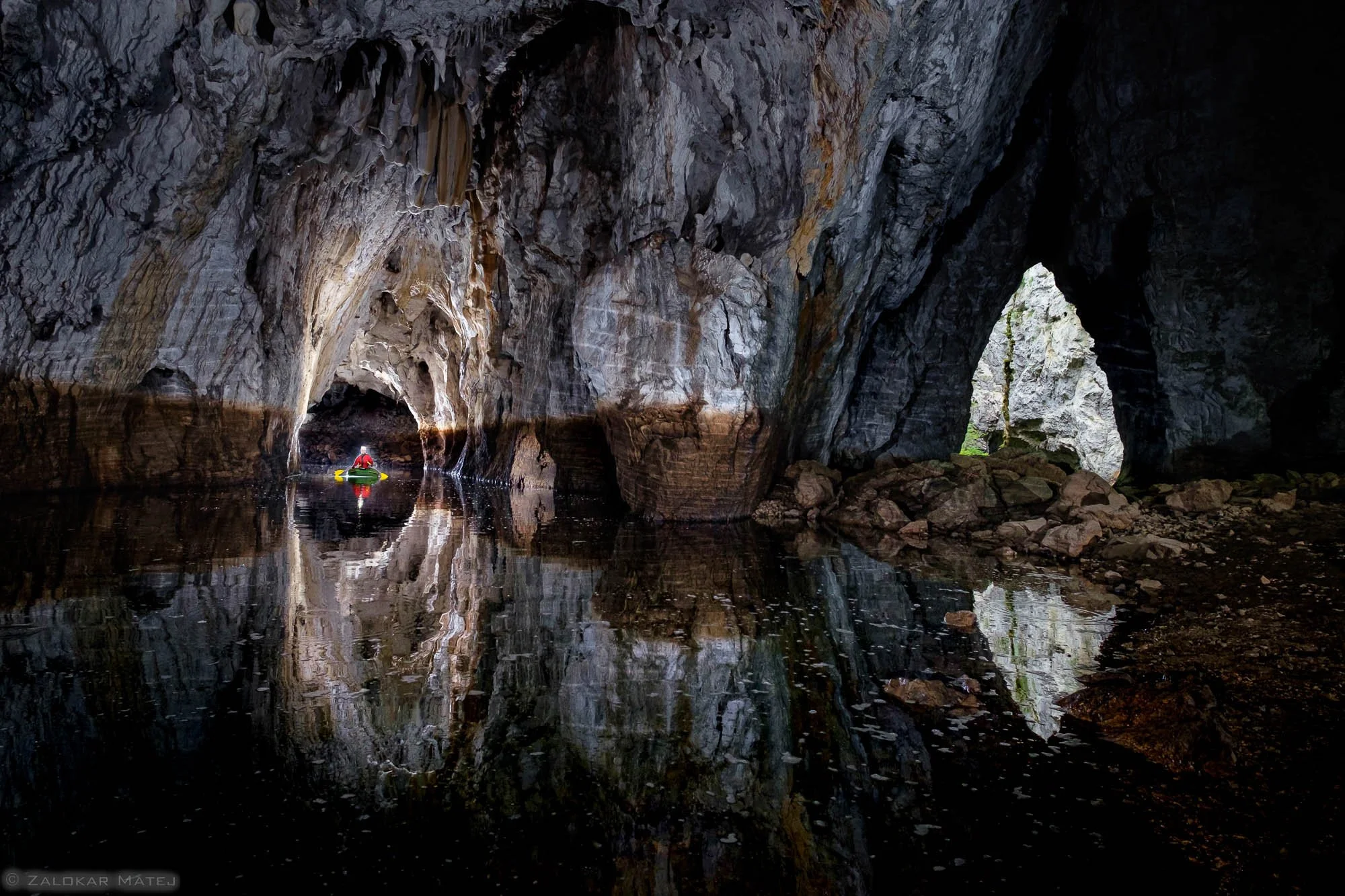 A person kayaking inside a large, dark cave with rocky walls and a water-filled floor, reflecting the cave's interior.