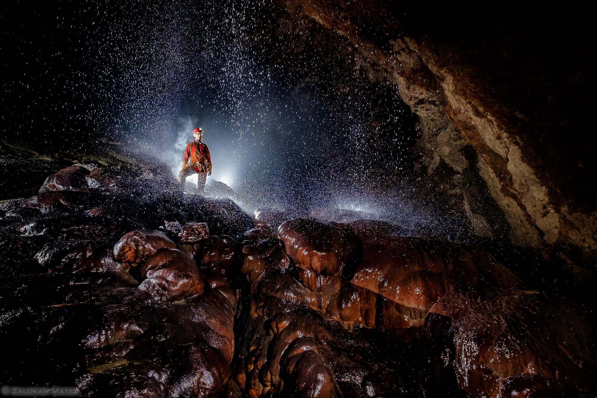 A caver standing on wet rocks inside a dark cave, spray of water arcing around him, illuminated by a light source behind.