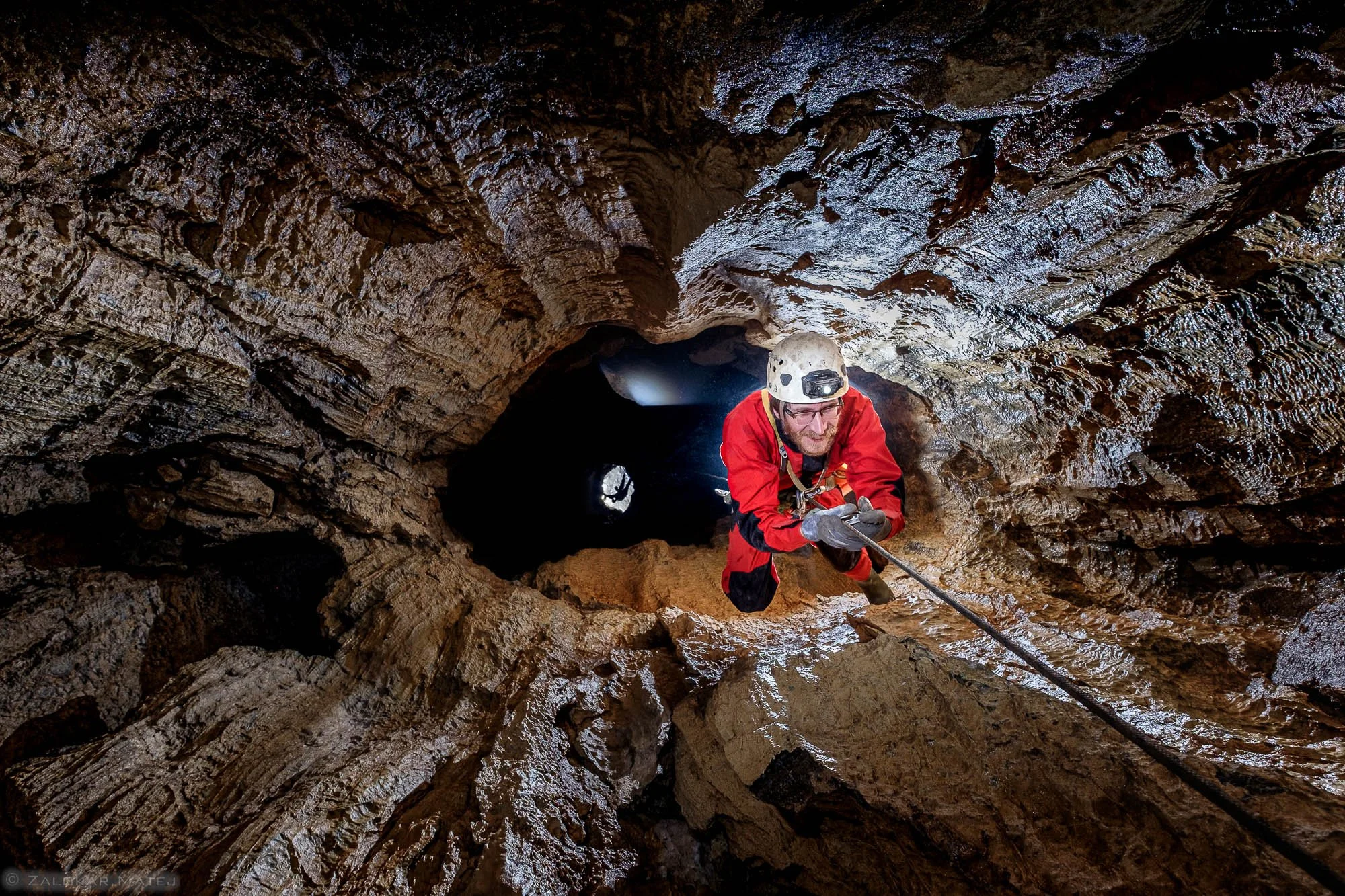 A man in a red jacket, helmet, and gloves rappels down the inside of a deep, narrow cave with rugged, wet rock walls.