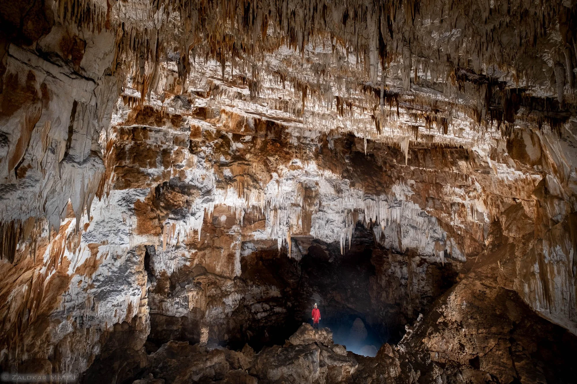 A person standing in a large cave with stalactites hanging from the ceiling and rocky formations on the ground.
