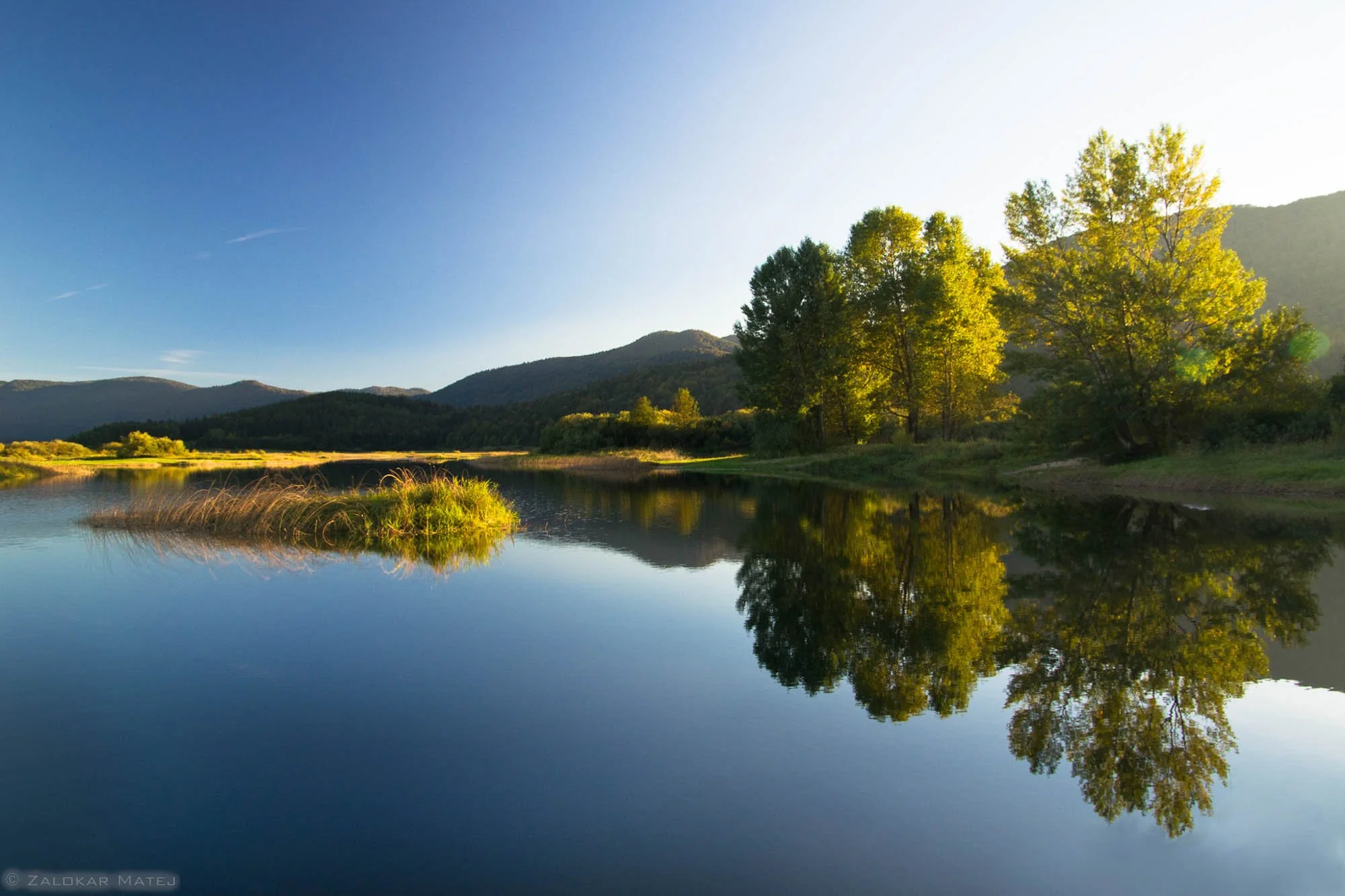 A serene landscape featuring a calm river reflecting trees and mountains in the background during late afternoon or early evening.