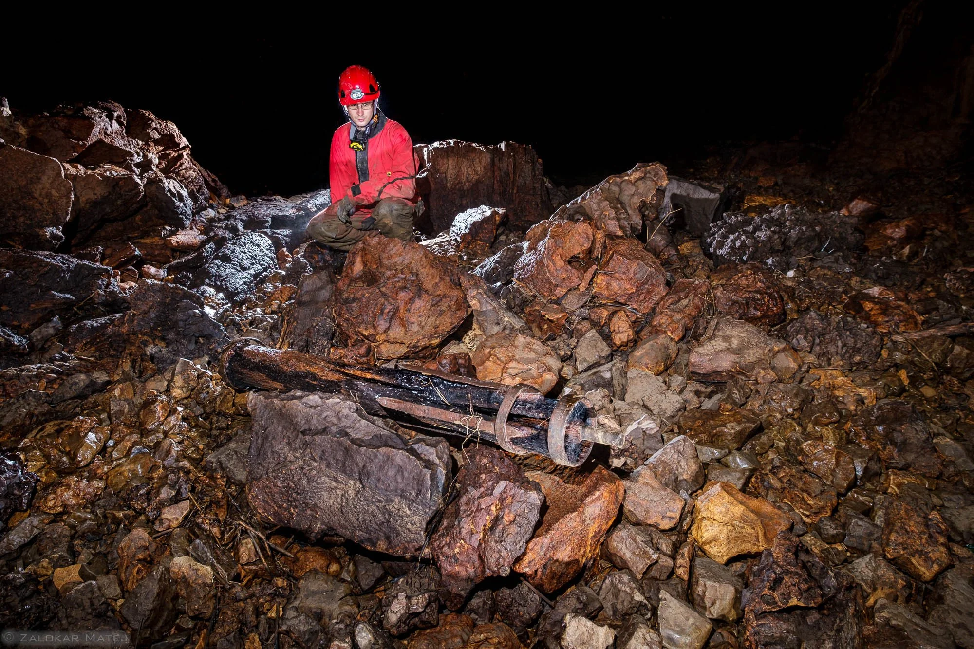 A person in protective gear and a red helmet kneels among large rocks inside a dark underground cave, with a drill-like tool lying on the rocks nearby.
