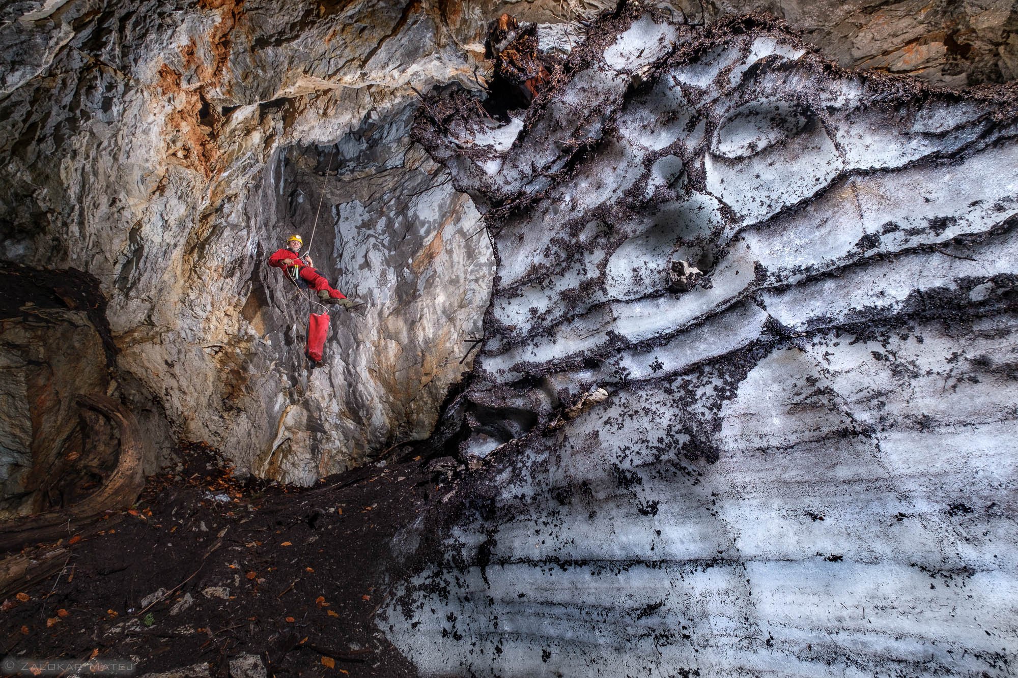 A person wearing red gear and a helmet climbing inside a rocky ice cave or crevasse, with icy and rocky surfaces overhead.
