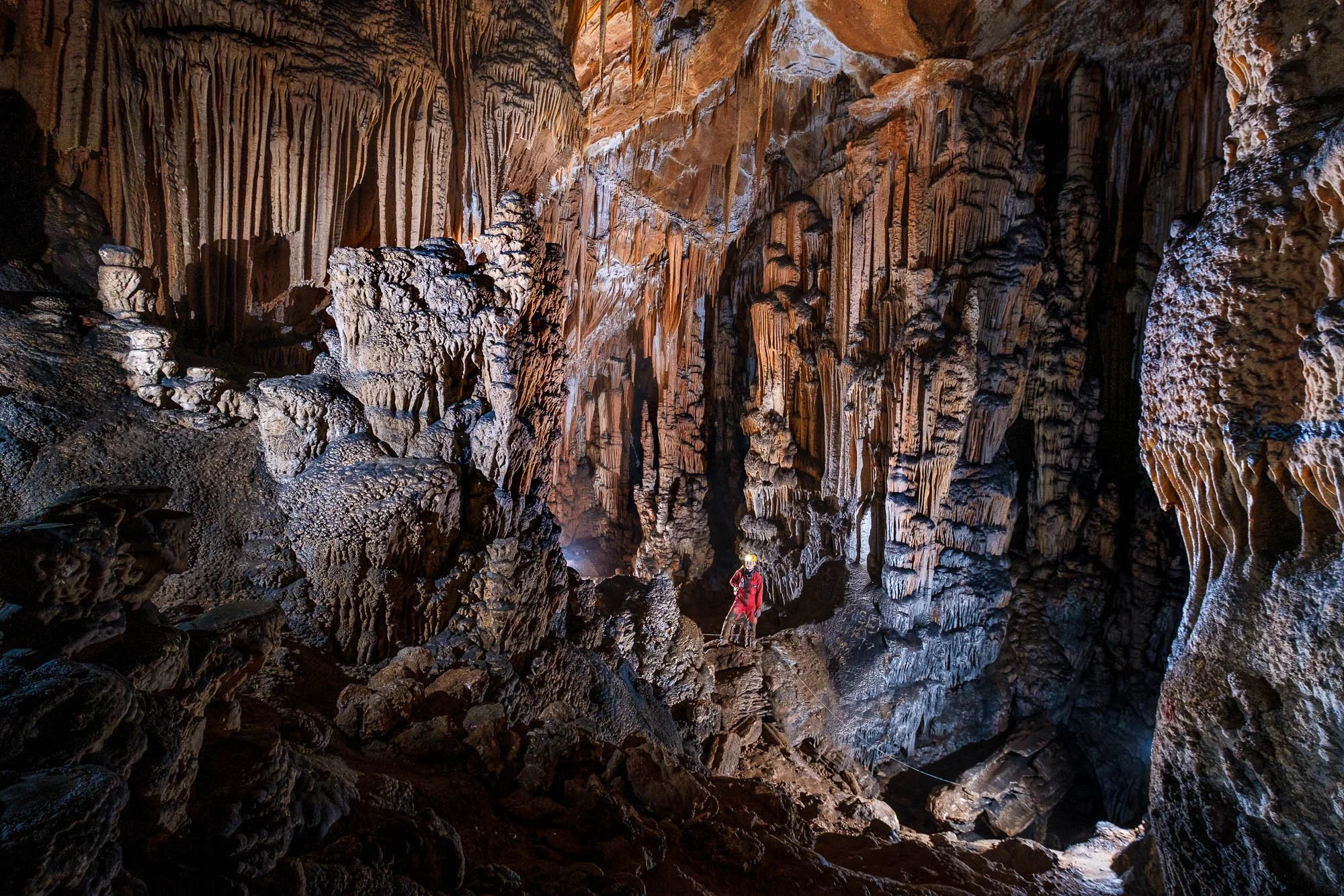 A person in a red jacket and helmet exploring inside a large cave with stalactites and stalagmites, illuminated by artificial lights.
