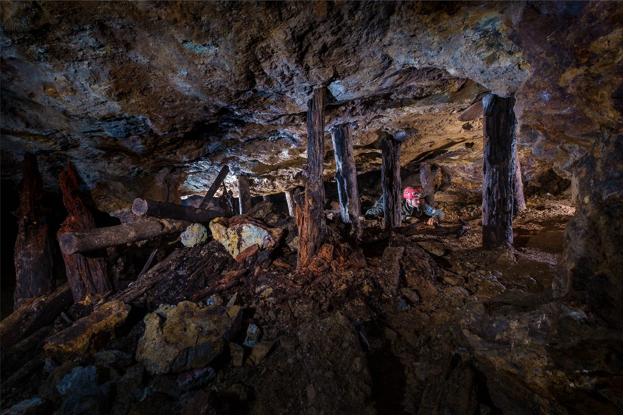 A person wearing a red helmet explores a dark, rocky underground cave with rough textured walls and ceiling, navigating over fallen logs and debris.
